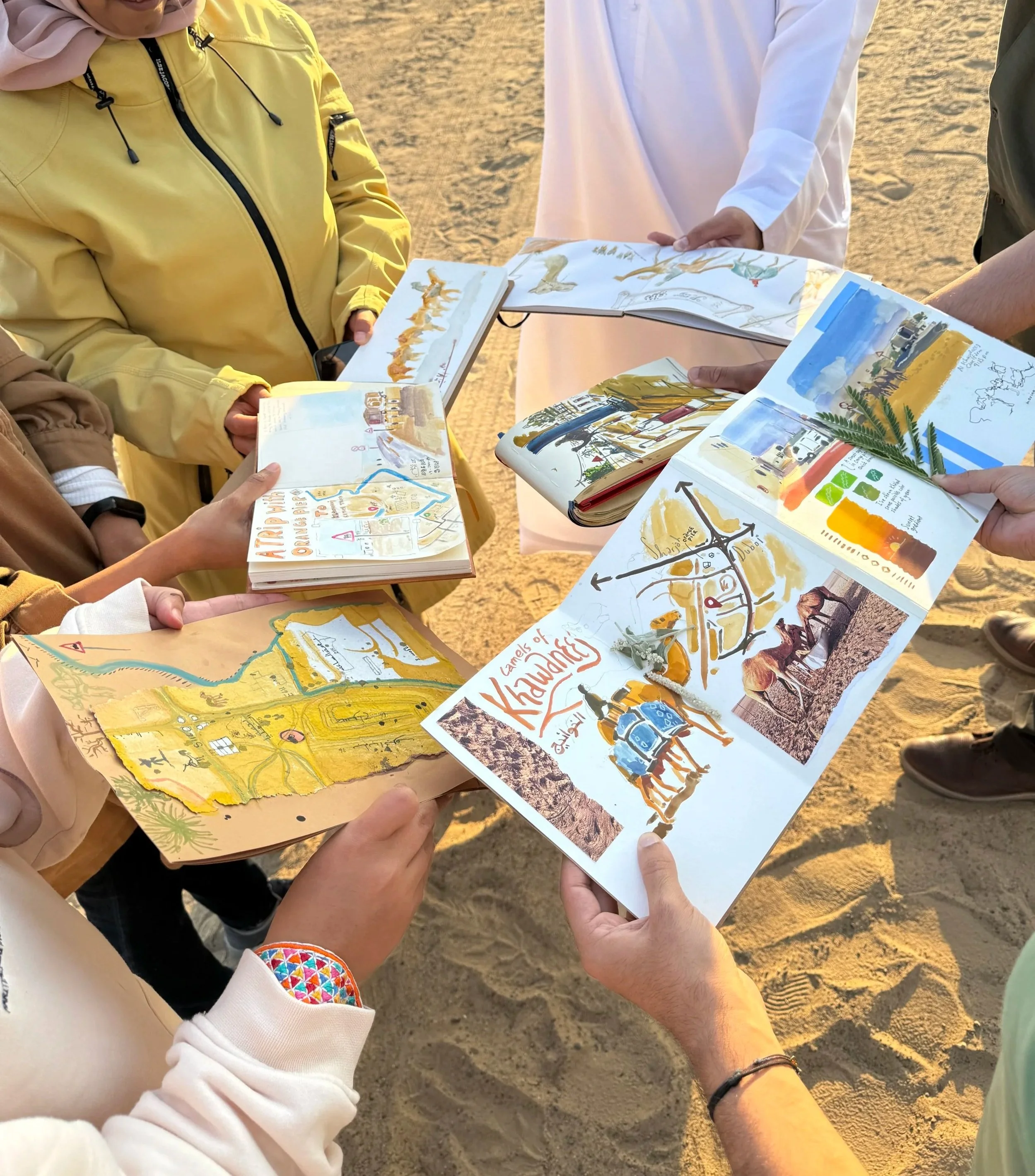 People holding colorful illustrated books or maps on a sandy outdoor area during sunset.