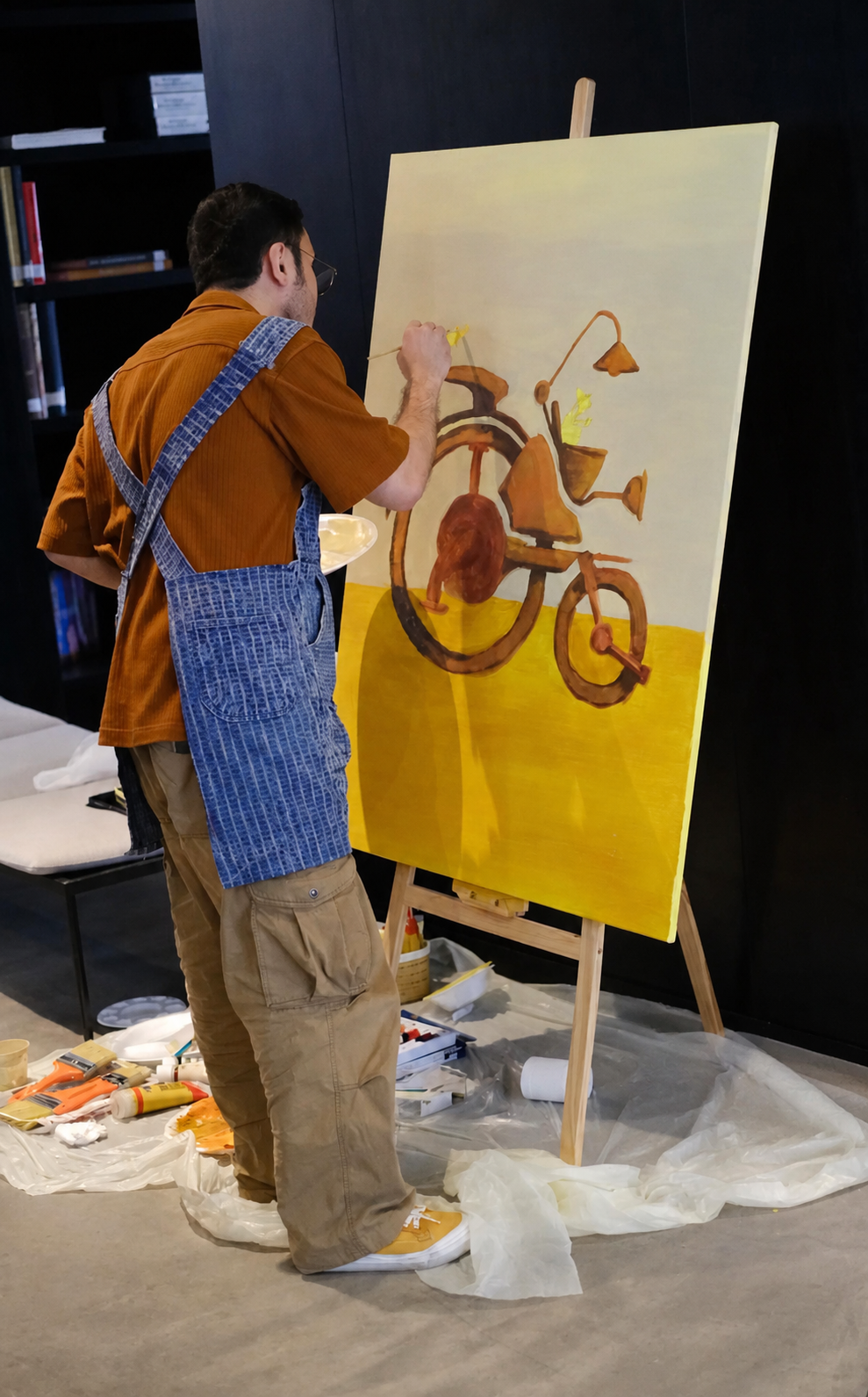 An artist painting a stylized bicycle on a large canvas, with paint supplies on the floor around him.