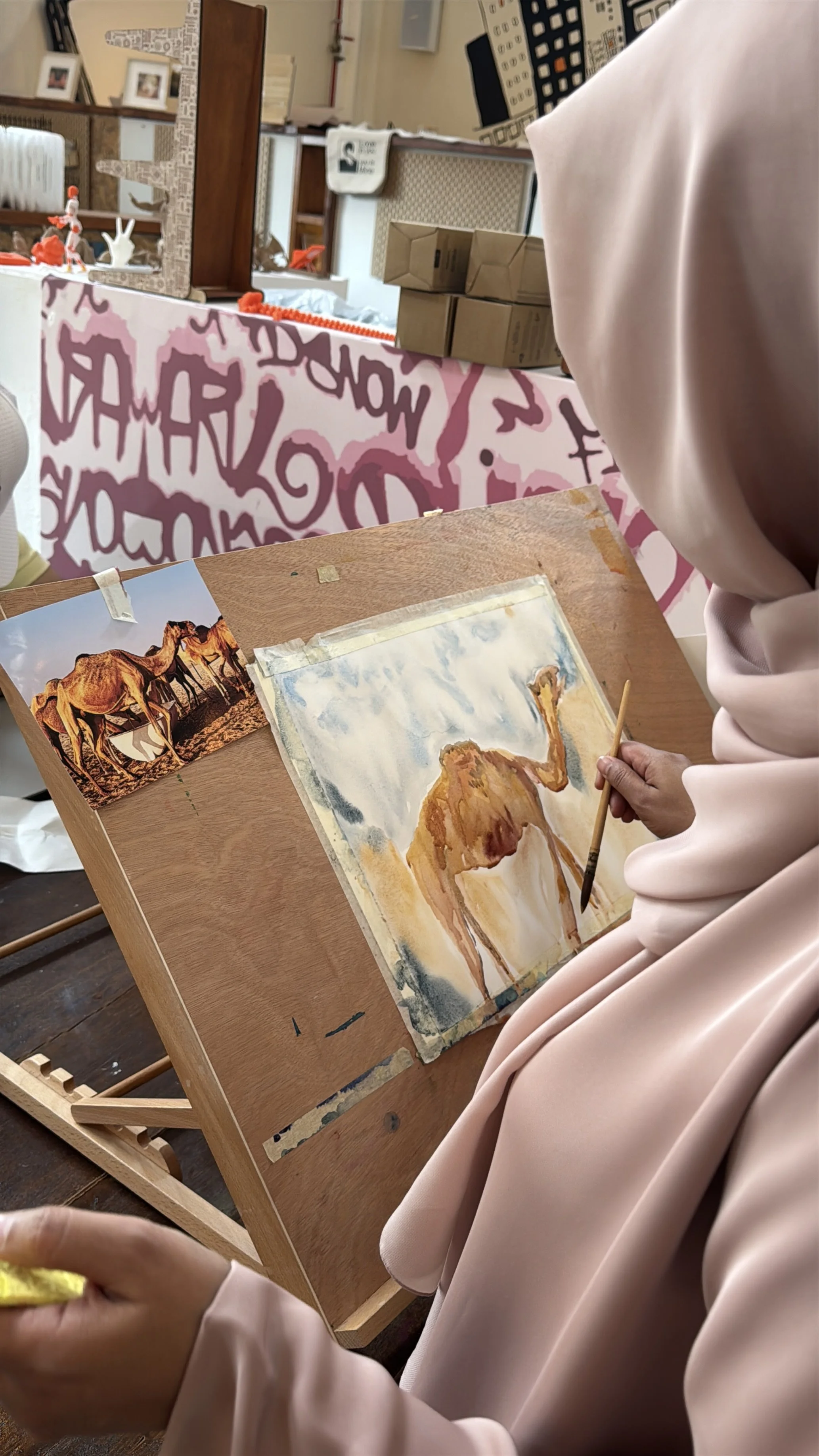 A person in a pink hijab painting a camel on a watercolor paper at an art event, with a printed photo of camels nearby.