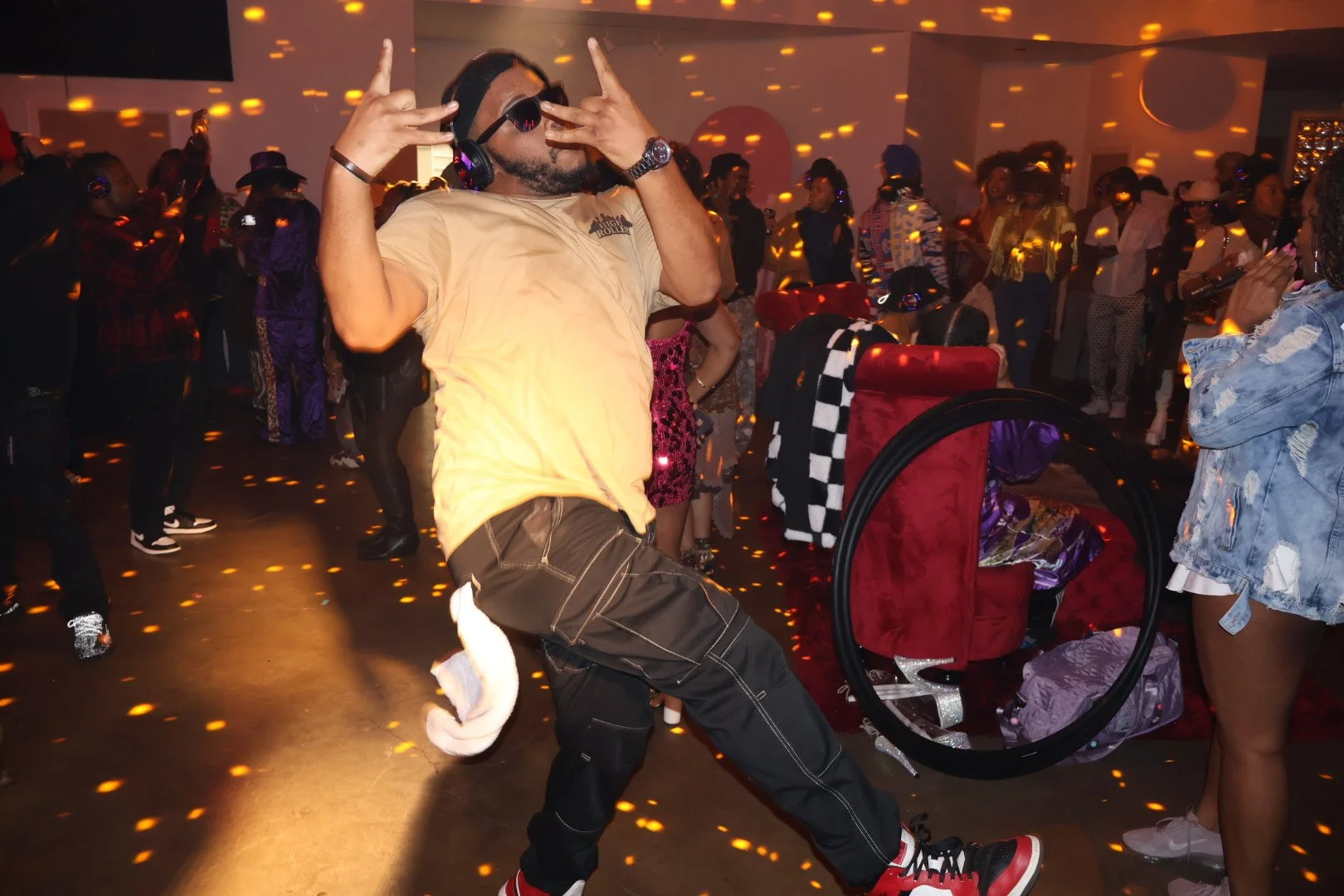 A man with sunglasses, headphones, a beige t-shirt, and dark pants is dancing with a playful pose in a lively indoor party. The background shows multiple people dancing and socializing, colorful lighting, and a festive atmosphere.