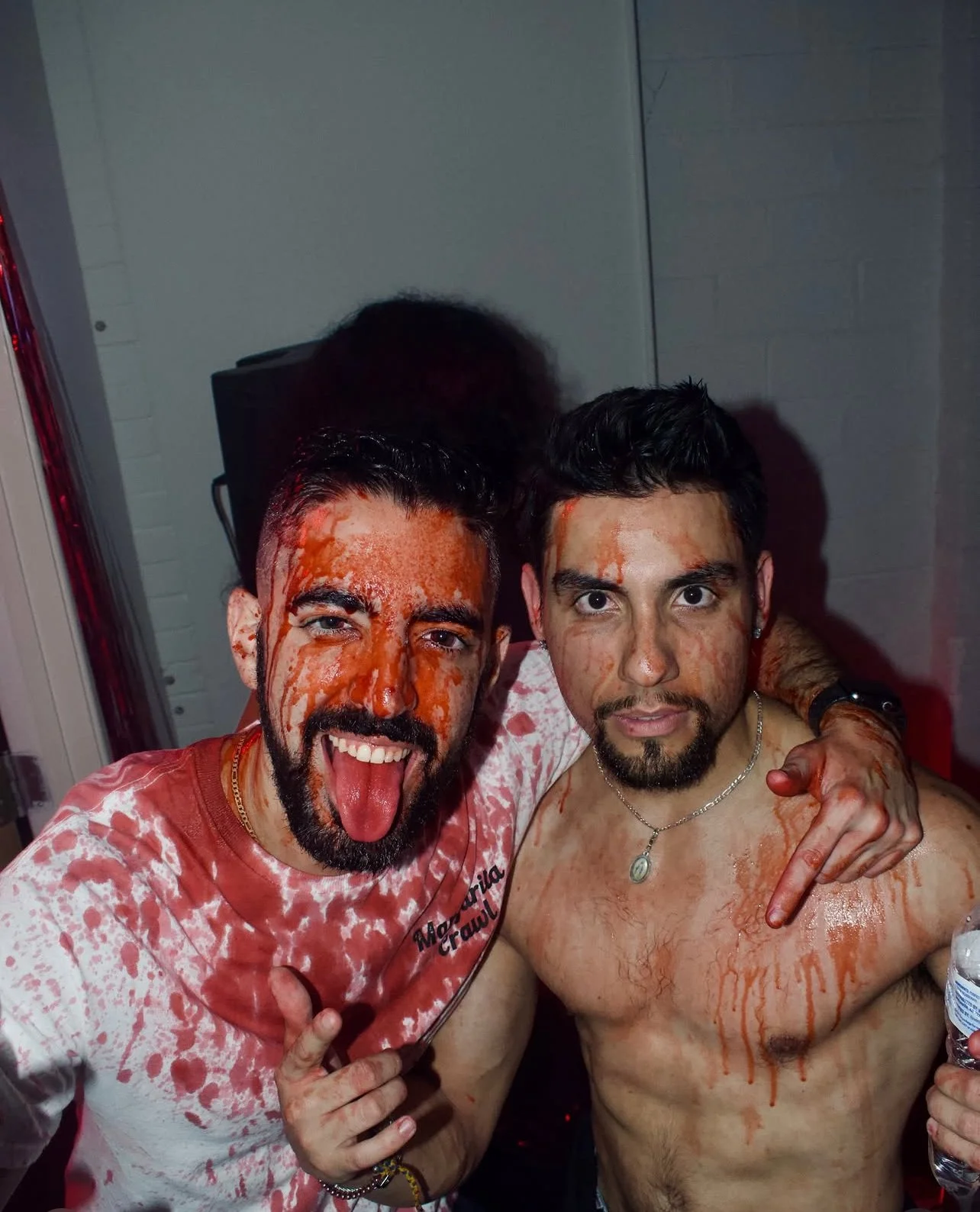 Two shirtless men with dark hair and facial hair celebrating, covered in red liquid, with one sticking out his tongue and the other making a hand gesture, indoors with a gray wall in the background.
