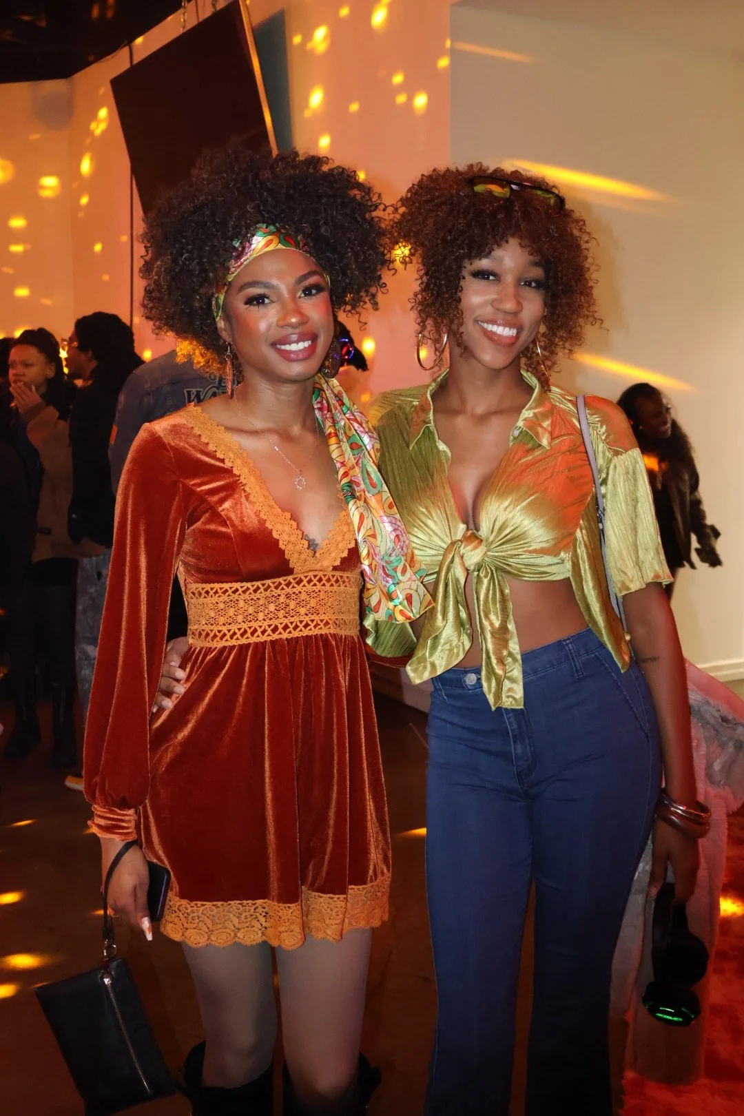 Two women with curly hair posing together at a party or event. One woman is wearing an orange velvet dress with lace details, the other is dressed in a shiny, golden-green tied blouse with blue jeans. The background shows people and warm lighting wit