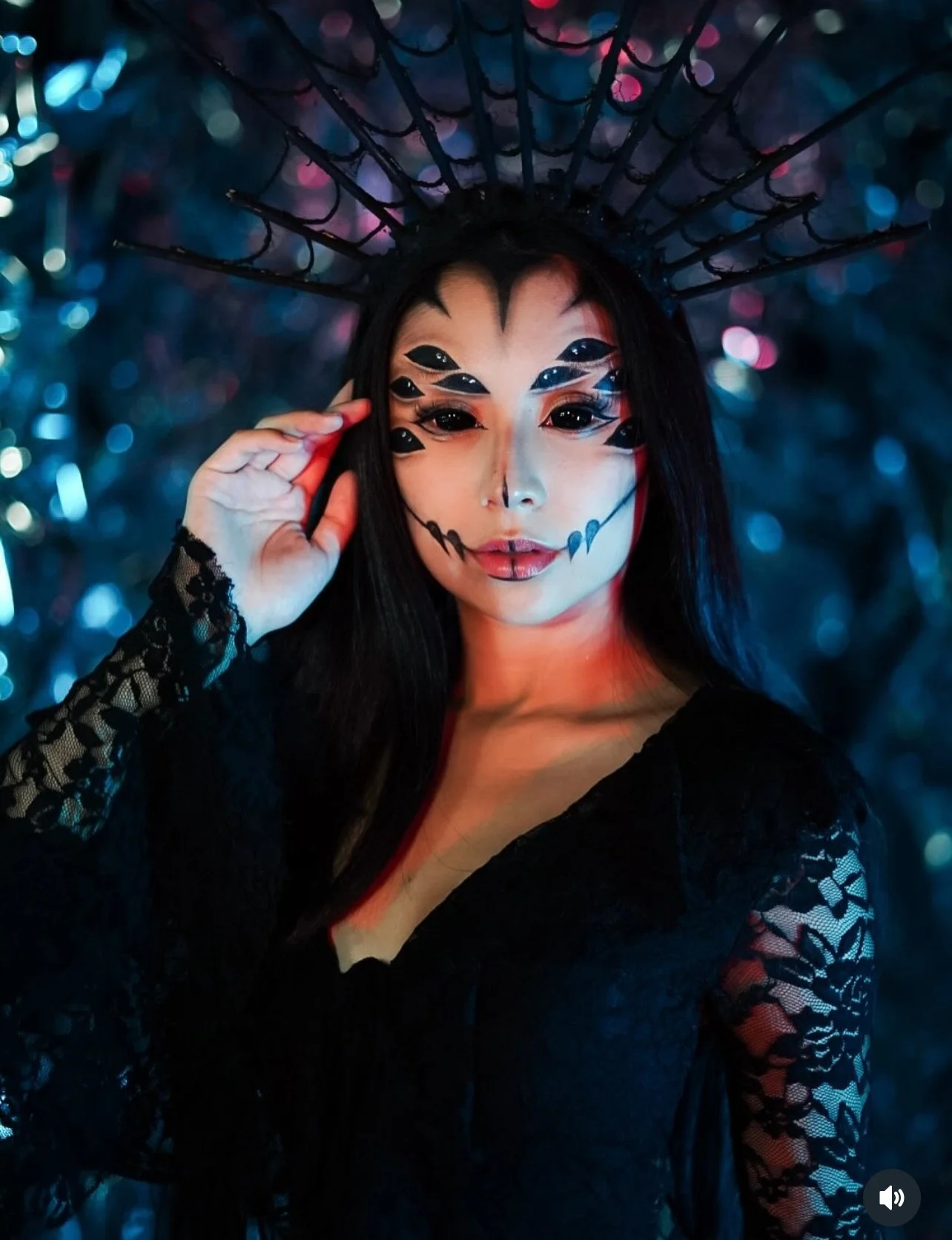Woman with face paint resembling a sugar skull, wearing black lace dress, black crown with spikes, and lace sleeves, with blurred colorful lights in the background.