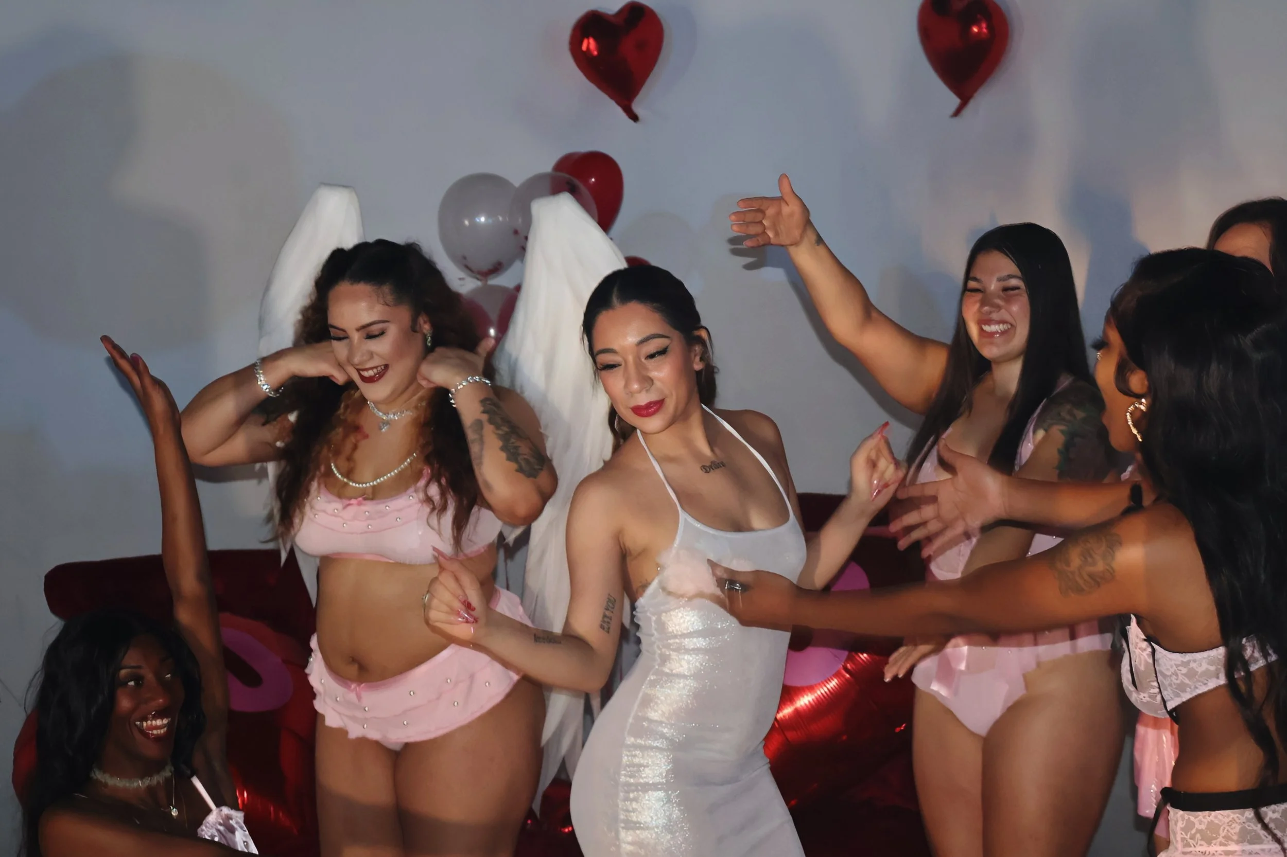 A group of women celebrating, some wearing pink lingerie, with balloons in the background, and a woman dressed as an angel with white wings at the center.