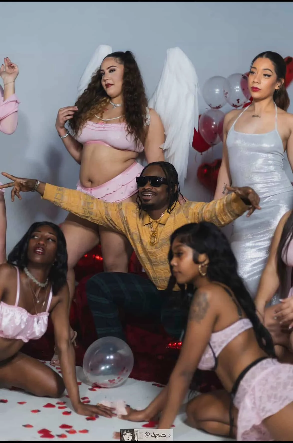 Group of people celebrating Valentine's Day with balloon decorations and scattered rose petals, including women in pink and white lingerie, a man in sunglasses, and angel wings worn by one woman.