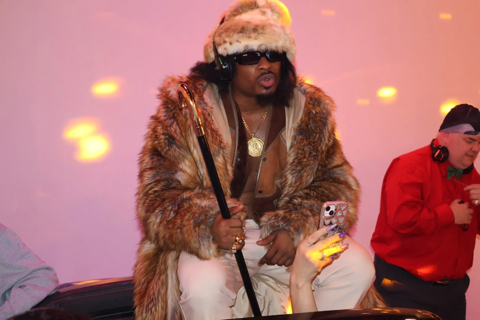 Man dressed in a fur coat with a gold medallion, hat, and sunglasses, holding a cane and a phone, sitting in a room decorated with pink and yellow lights, with another person in red in the background.