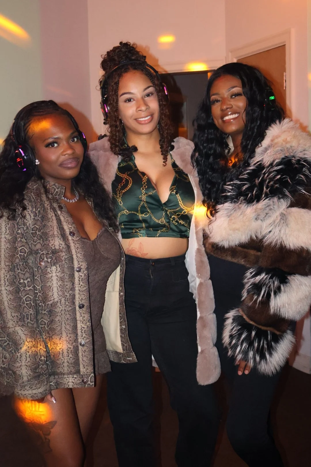 Three women posing together at an indoor event, wearing stylish outfits and headphones, with warm lighting and a neutral background.
