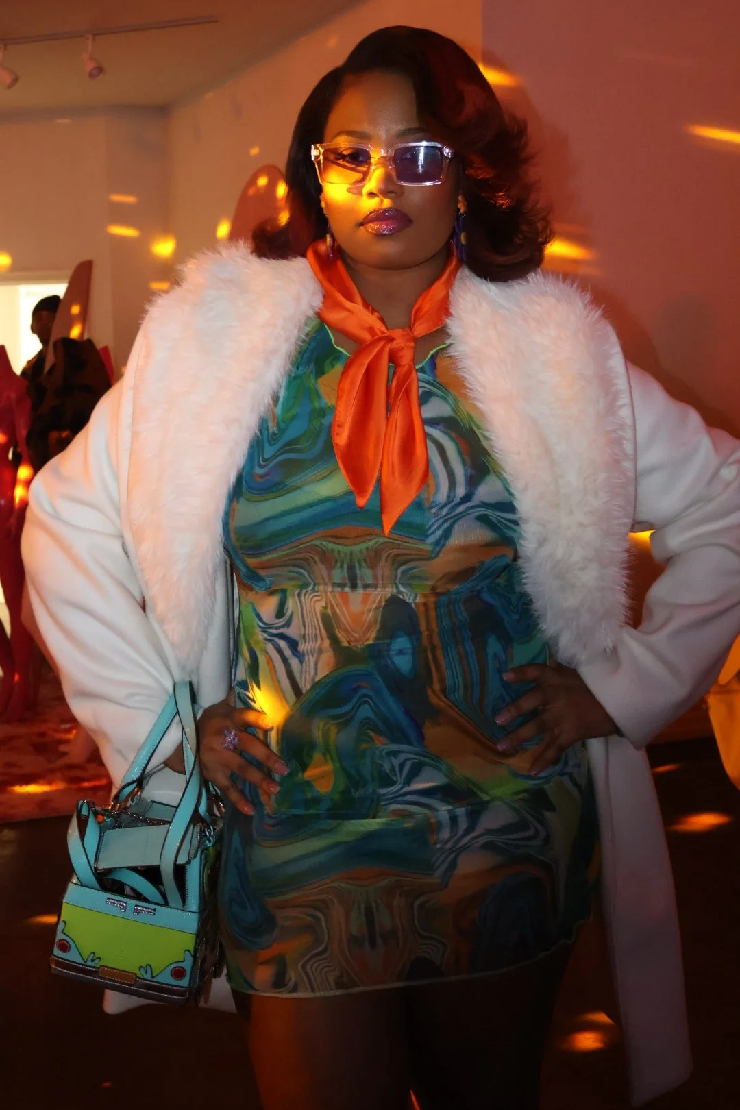 A woman with dark hair styled in loose curls, wearing sunglasses, a colorful swirl-patterned dress, a white fur-lined coat, and an orange scarf tied around her neck. She has a small handbag with a cartoon design hanging from her arm and is standing w