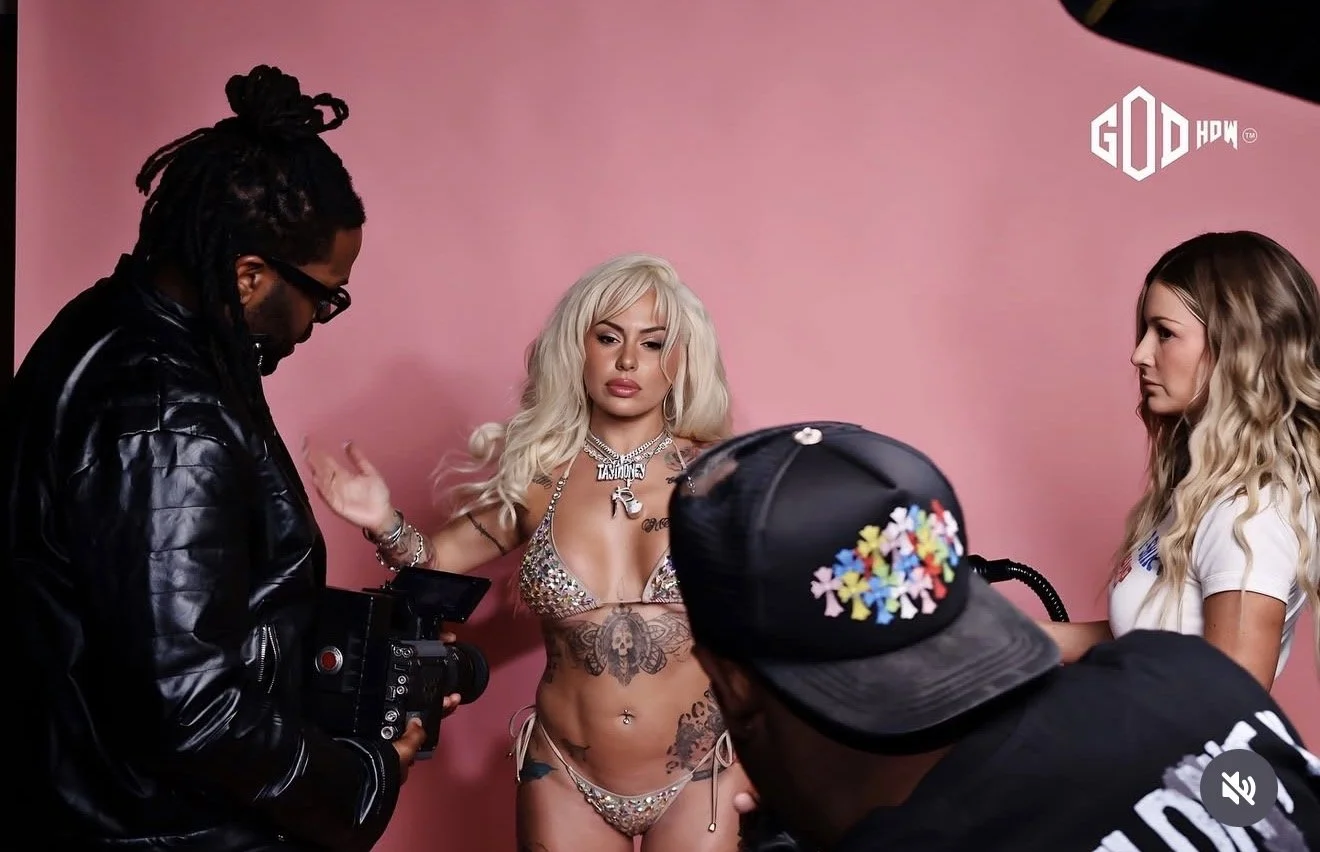 A woman with long blonde hair and tattoos wears a sparkly bikini, being photographed/filmed with a pink background and crew members around her.