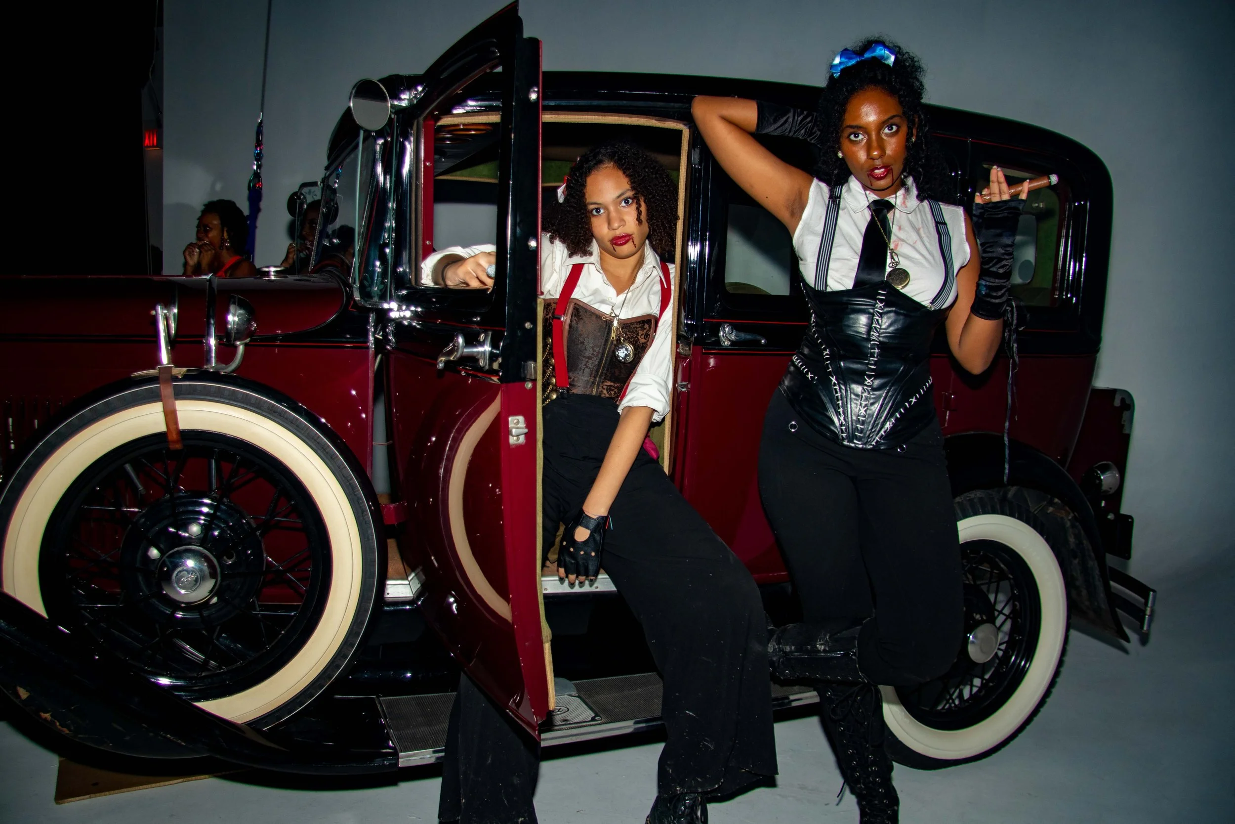 Two women in vintage-inspired outfits and makeup posing with a classic red and black automobile inside a studio.