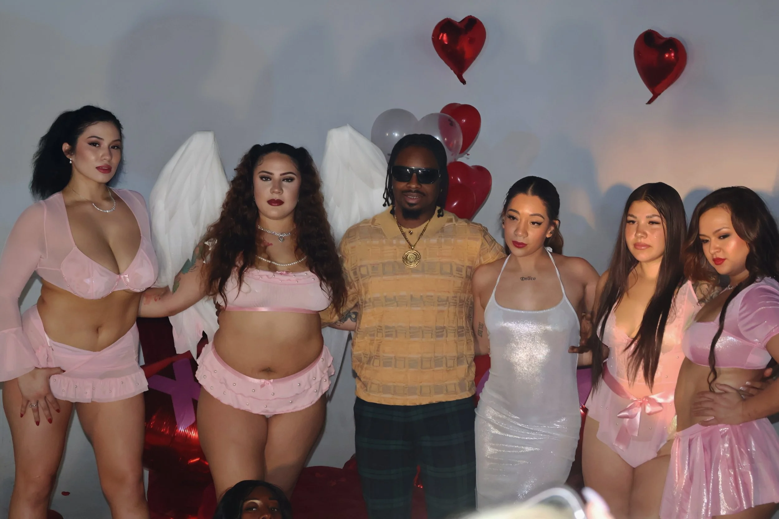 Group of six people standing together with red and white balloons and heart-shaped balloons in the background.
