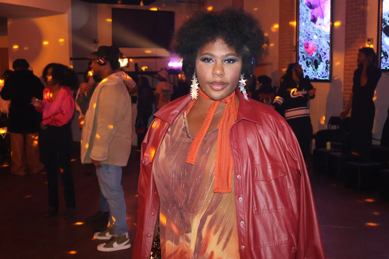 A woman with short curly black hair wears a red leather jacket over a sheer, orange patterned top, standing in a dimly lit lounge or club with other people in the background. There are bright digital screens on the walls and colorful lighting effects