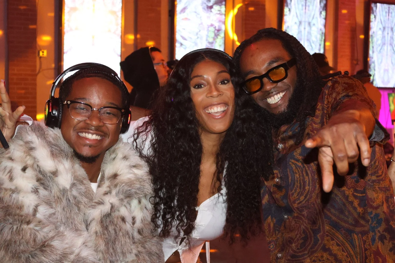 Three people, two men and one woman, smiling and enjoying a lively indoor event. The man on the left is wearing glasses, headphones, and a fur coat. The woman in the middle has long curly black hair and is dressed in a white top. The man on the right