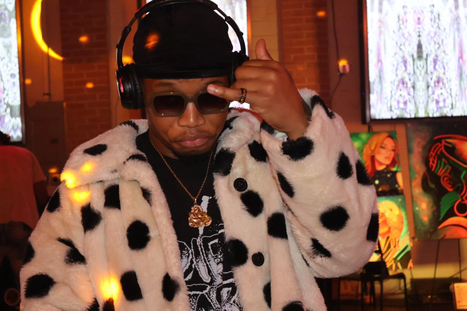 A person wearing sunglasses, headphones, a black cap, a black shirt with a skeleton design, a gold necklace, and a white faux fur coat with black spots, standing in an indoor setting with artwork and illuminated displays in the background.