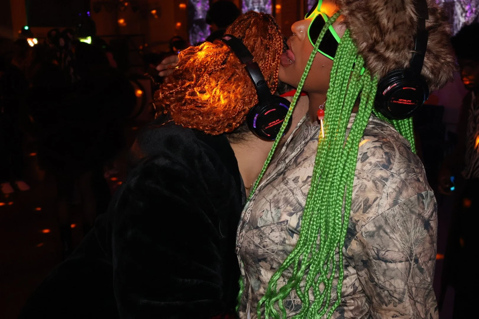Two people with vibrant hair colors and large headphones sharing a kiss at a party, with one having orange hair and the other green braided hair, wearing a camouflage jacket and black velvet top.