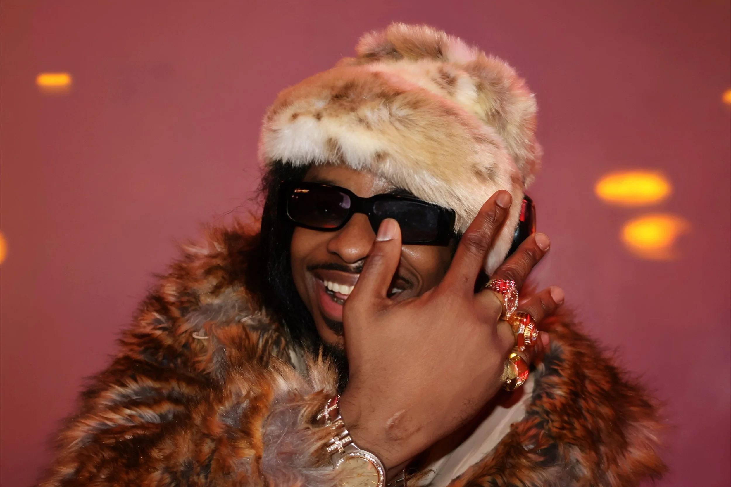 A person wearing a fluffy, patterned fur coat, dark sunglasses, and multiple gold rings, adjusting their sunglasses with a smile against a pink background with blurred orange lights.