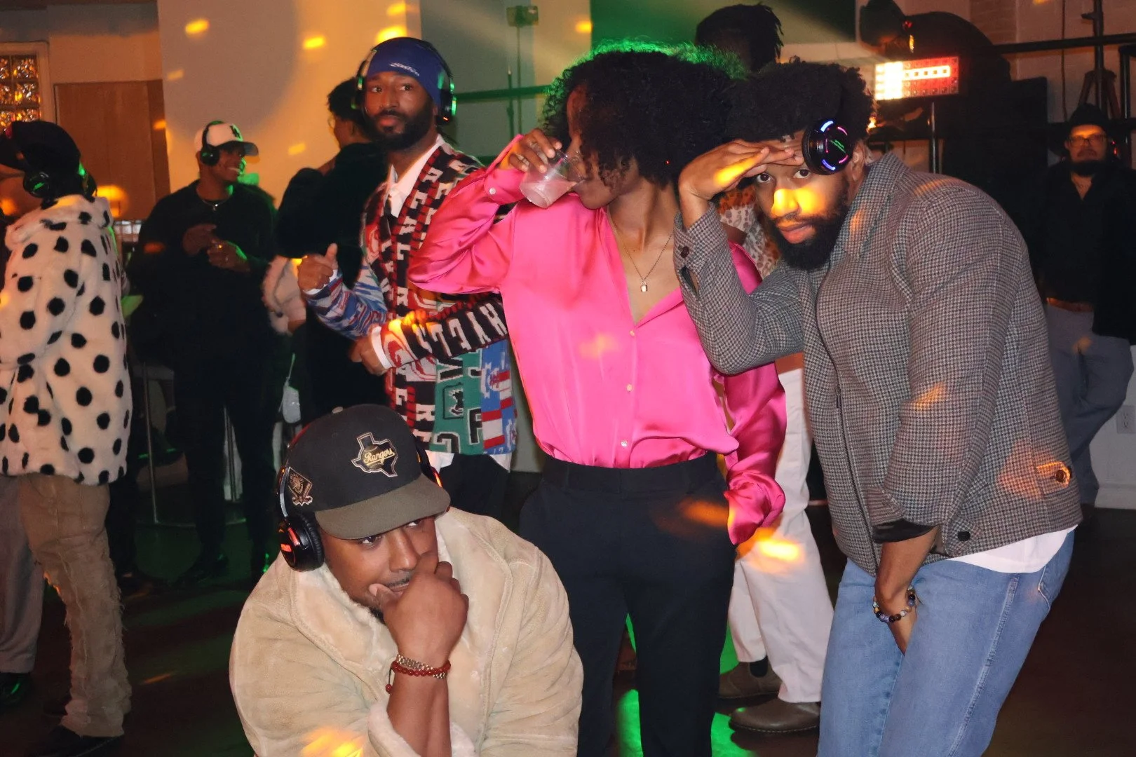 People dancing and socializing at a party with colorful lighting, including a woman in a pink satin shirt and a man with headphones and a checkered blazer, surrounded by others dressed casually.