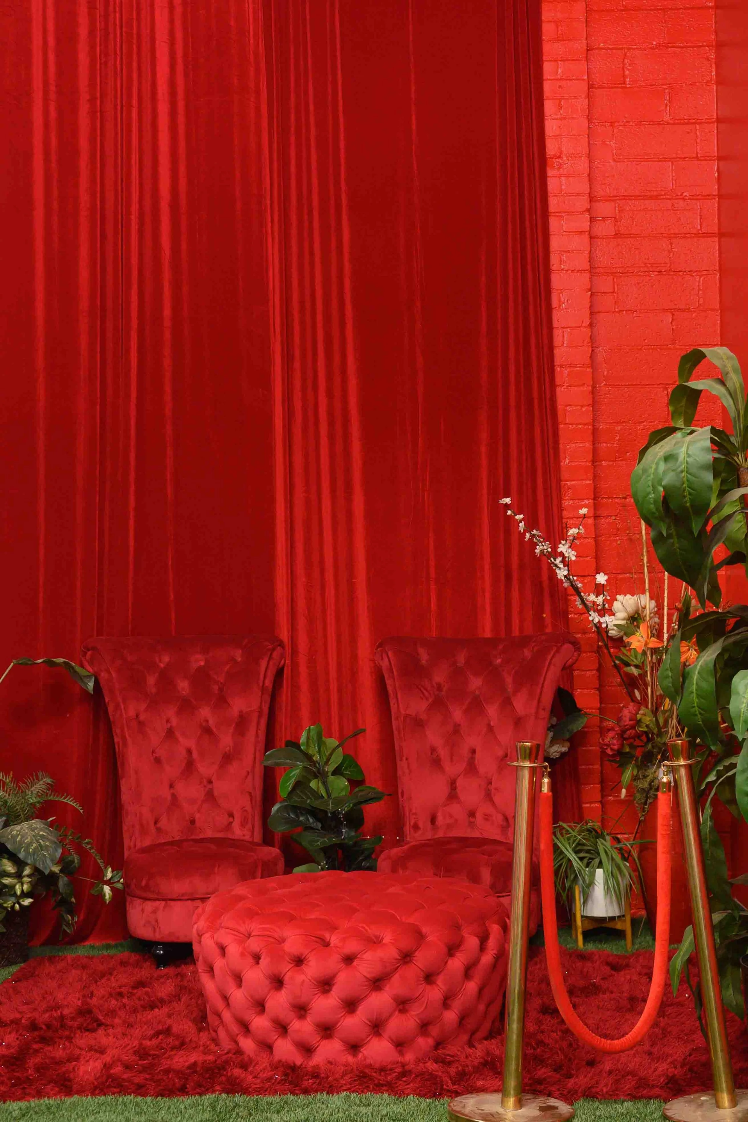 Red velvet chairs with tufted backs and matching ottoman in front, set against a red brick and velvet curtain background surrounded by green plants and flowers.