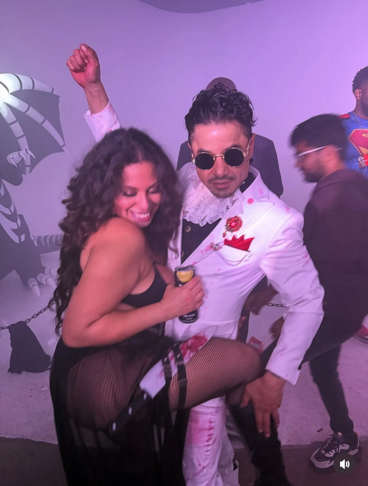 People at a party pose for a photo, with a woman smiling and a man in a white suit wearing sunglasses holding a drink. Other party guests are in the background.