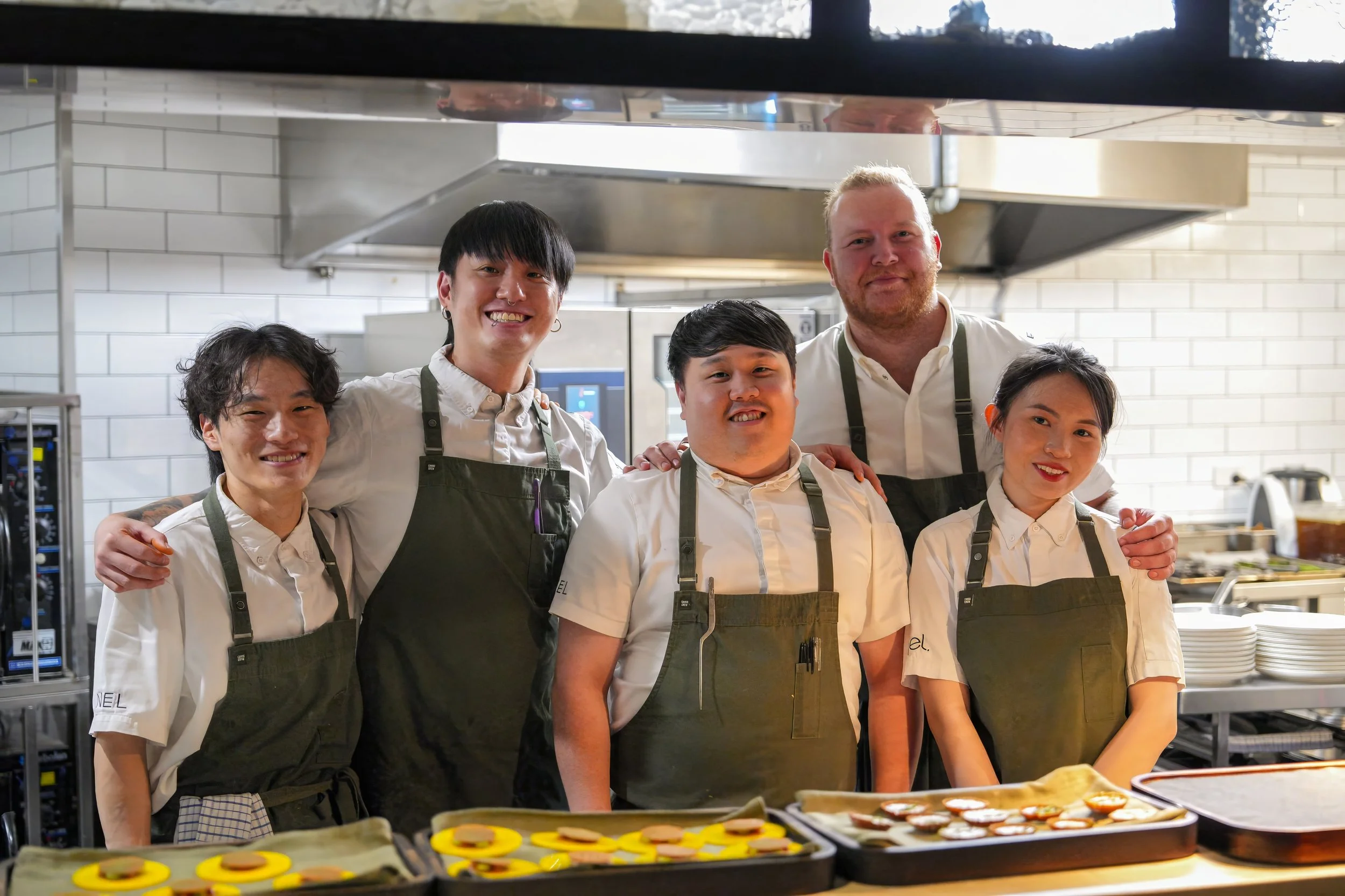 Group of five chefs in a commercial kitchen smiling at the camera, with trays of food in the foreground. Sydney. Studios Metaversal.