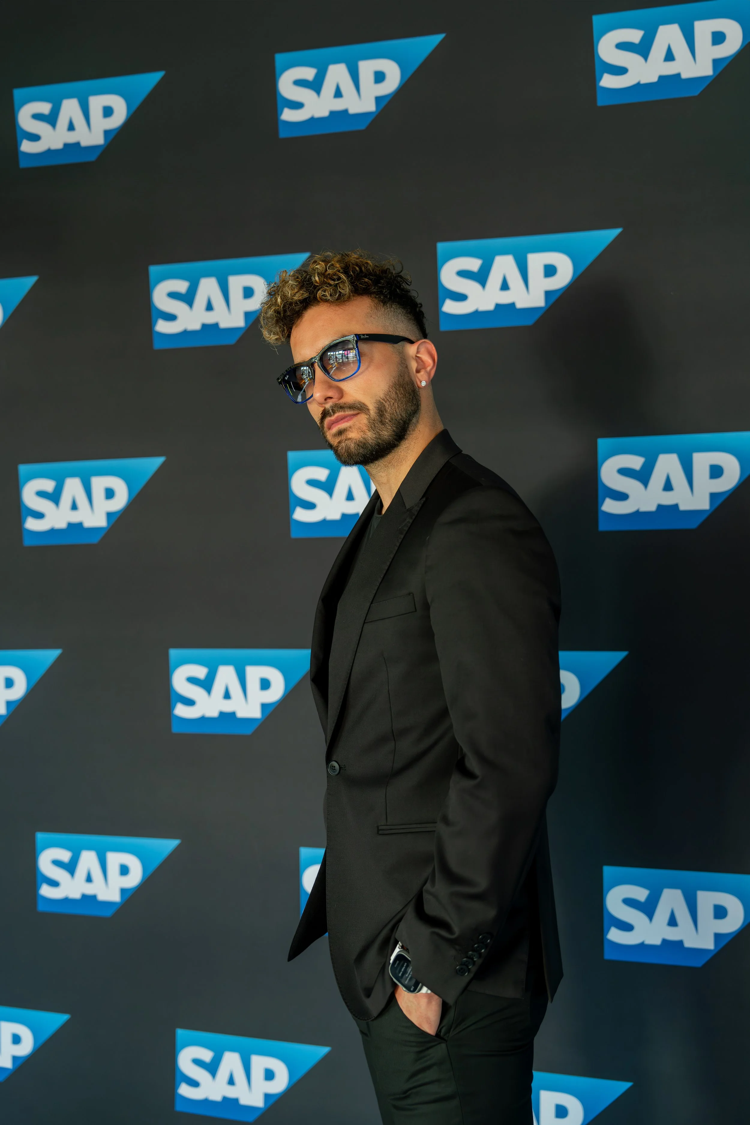 A man with curly hair, beard, and sunglasses, wearing a black suit, posing against a backdrop with the SAP logo. Javo El Cirujano. Studios Metaversal