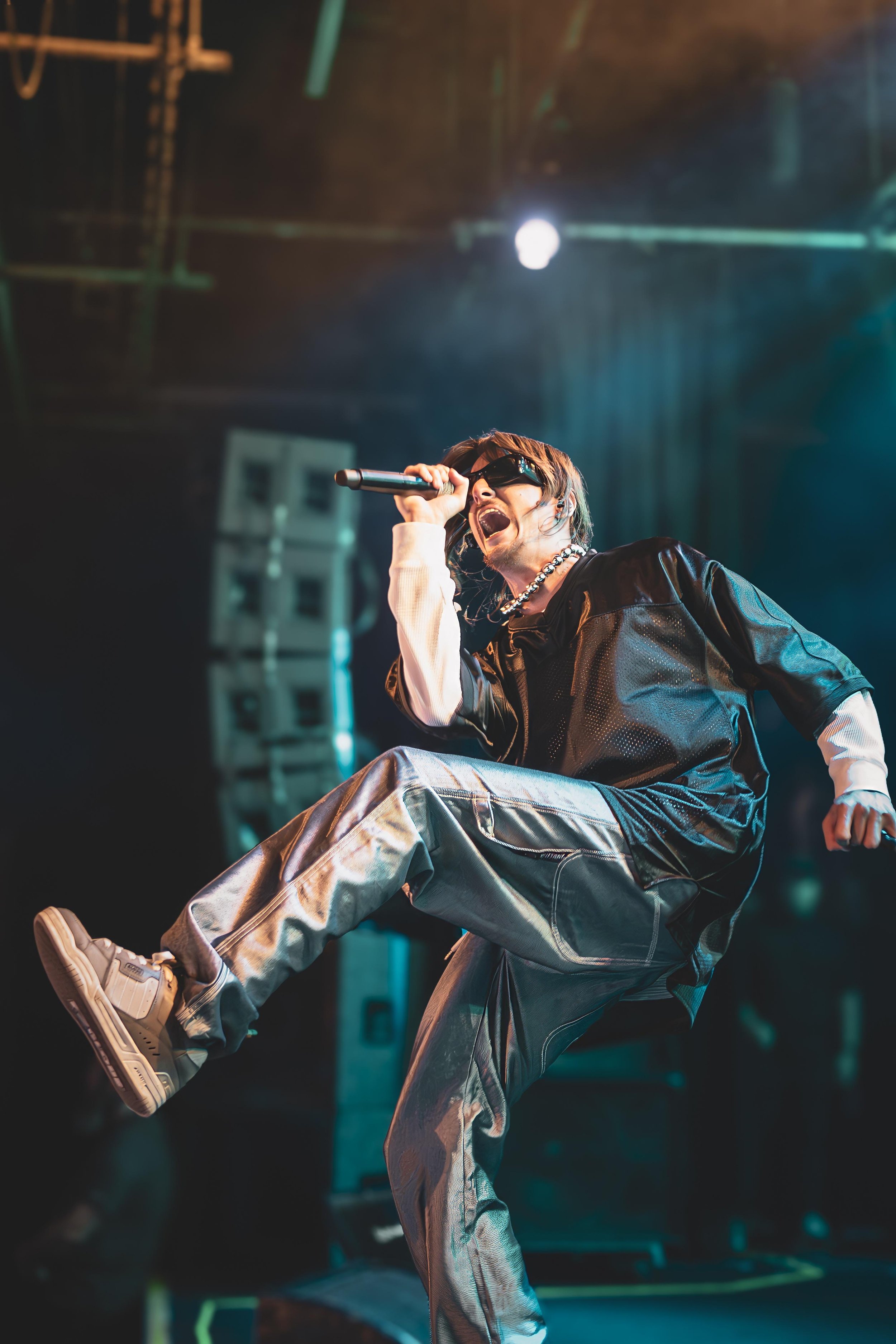 Performer singing into microphone on stage, wearing sunglasses, a black leather jacket, and metallic pants, with energetic pose.