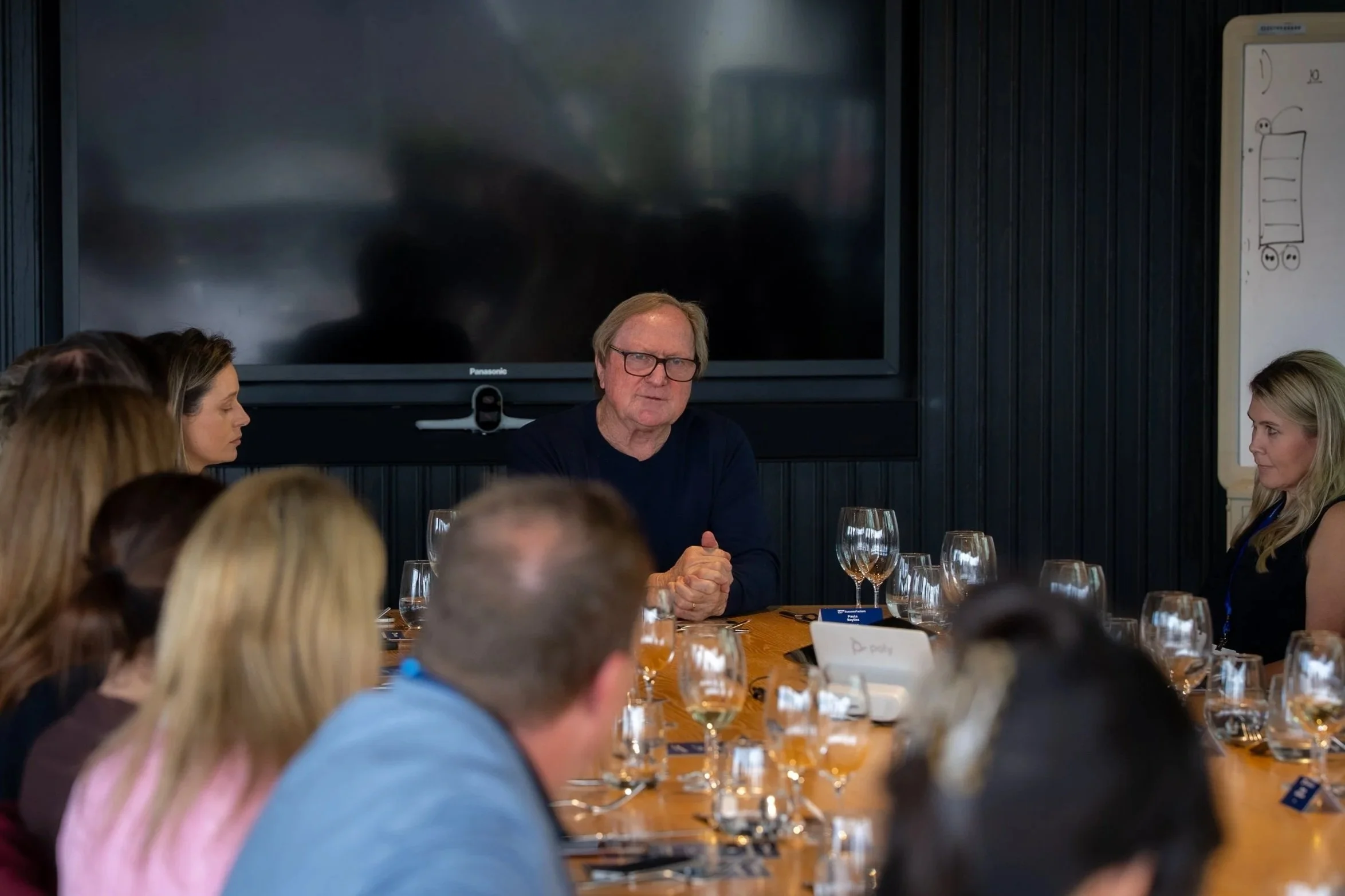 A group of people sitting at a long dining table in a conference room, with a man with glasses and a dark shirt speaking in front of a large blank screen. Melbourne. Studios Metaversal