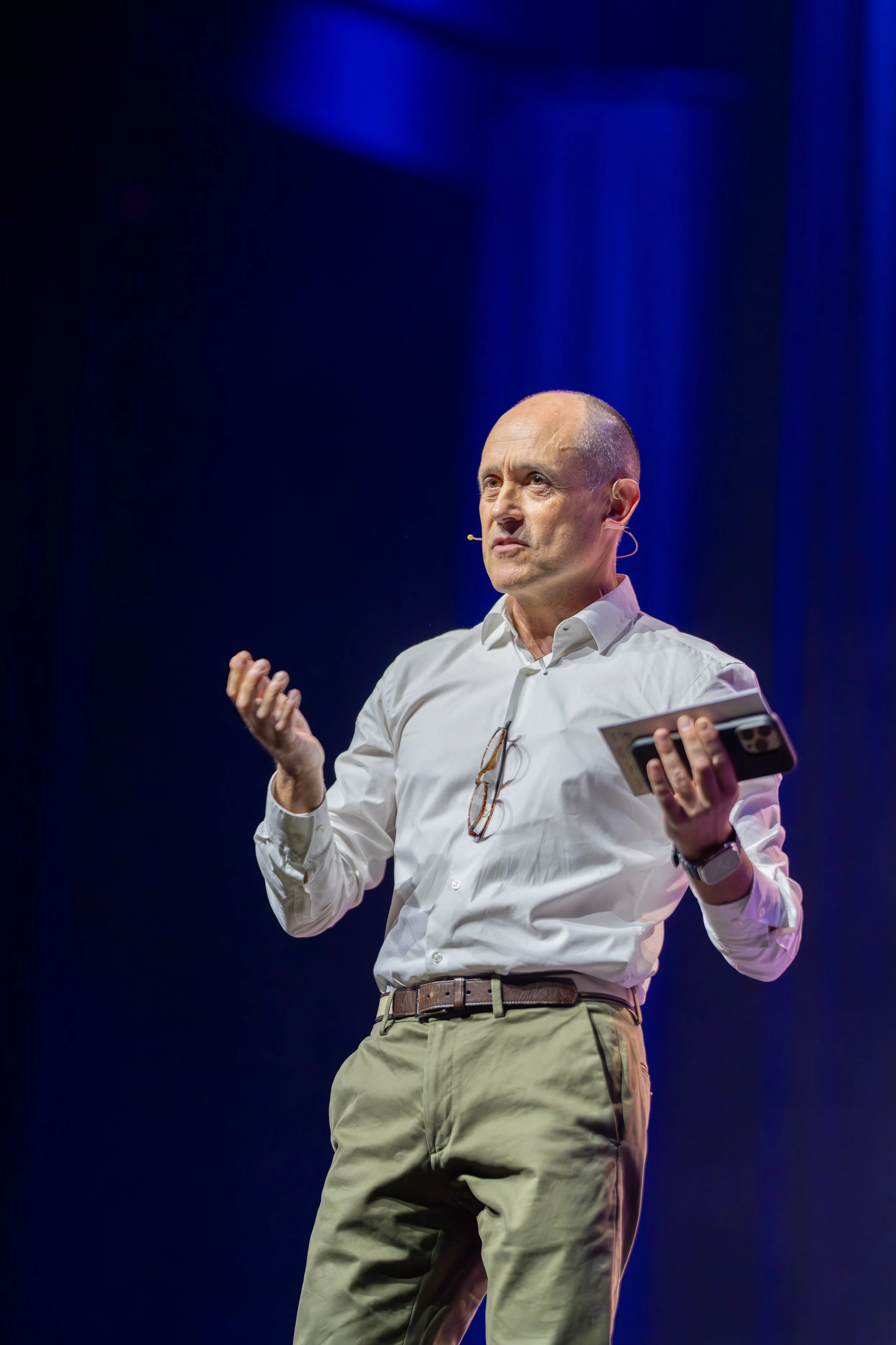 A bald man wearing a white shirt, beige pants, and a watch, speaking on stage with a microphone headset, holding a device, against a dark background with blue lighting.