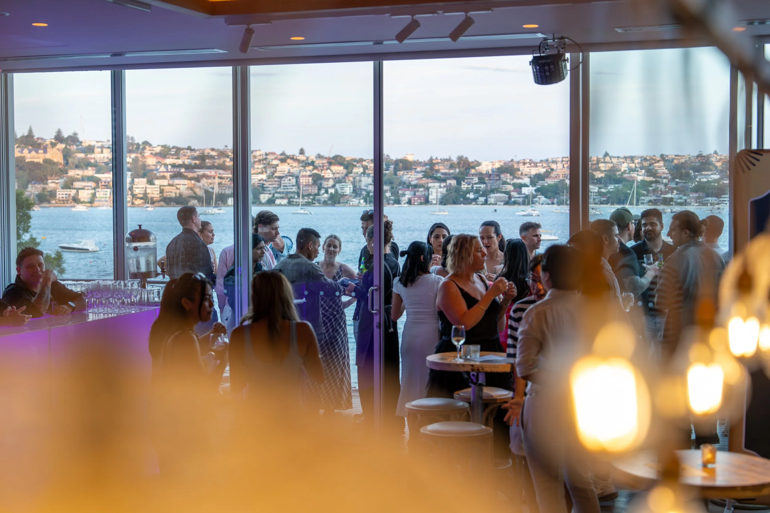 People gathered inside a restaurant with large windows overlooking a waterfront with boats and hillside houses. Sydney. Studios Metaversal.