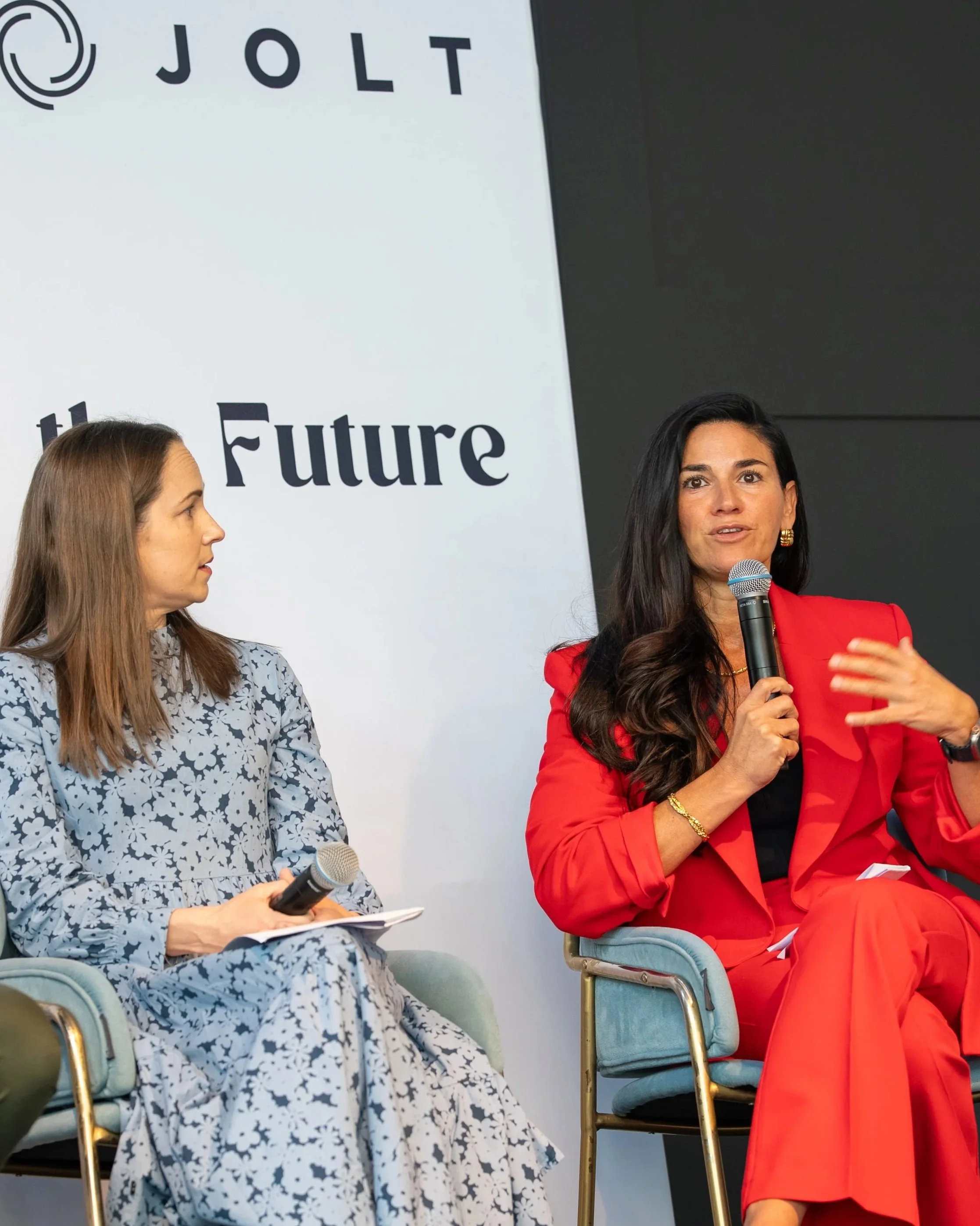 Two women seated on stage at a panel discussion. The woman on the left has long brown hair, is wearing a blue dress with a floral pattern, and holds a microphone and notebook. The woman on the right has long dark hair, is wearing a red blazer and matching skirt, and is speaking into a microphone. A large white screen behind them displays text that reads 'JOLT' and 'the Future'.