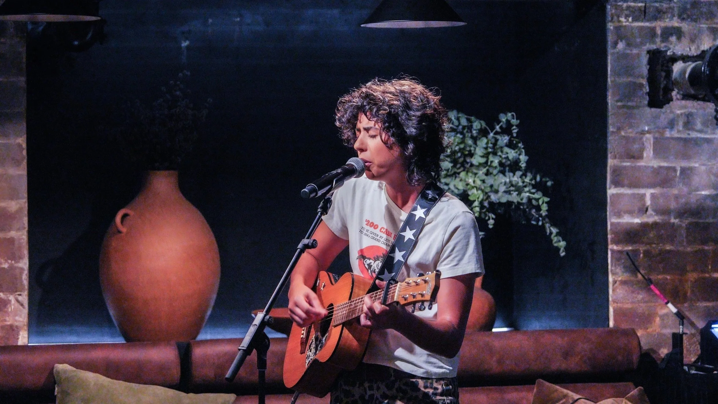 A woman with curly hair singing into a microphone while playing an acoustic guitar in a dimly lit venue with brick walls and a decorative vase in the background. Sydney. Studios Metaversal.