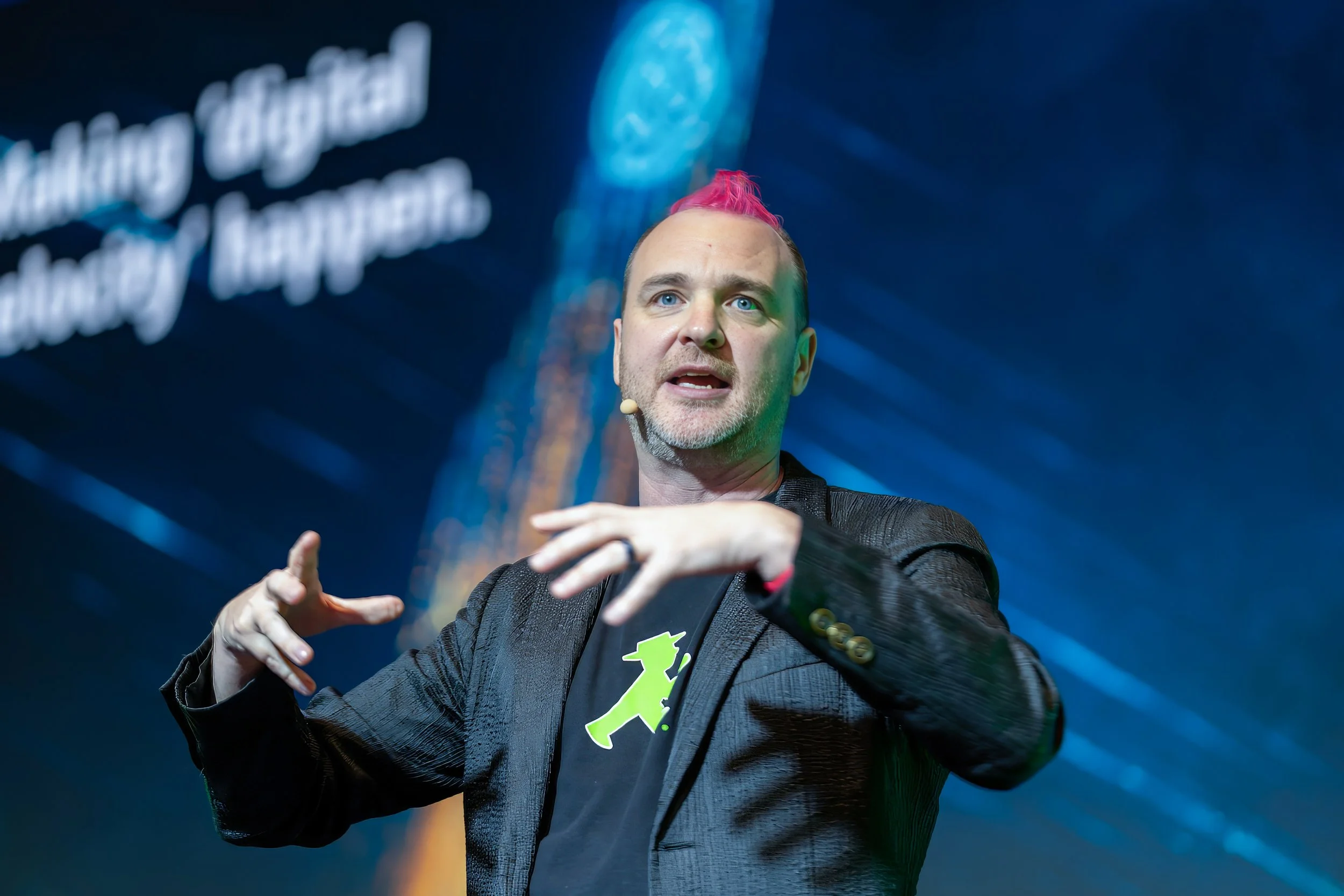 A man with pink hair, a beard, and blue eyes speaking at an event with a dark background. He is wearing a black blazer over a shirt with a neon green and black graphic. His hand gestures are expressive, and he is wearing a small microphone on his che