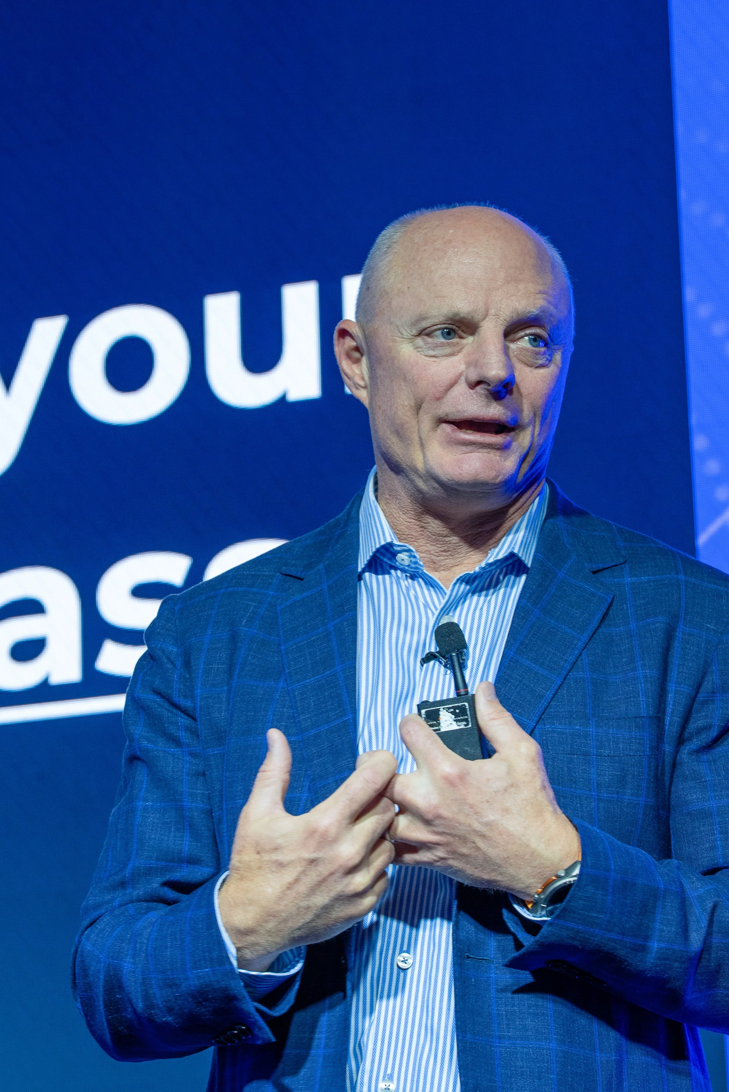 A man with a bald head and light blue eyes speaking at a conference or event, wearing a blue checkered suit and blue striped shirt, gesturing with his hands, with a microphone clipped on his shirt, against a blue background with white text.