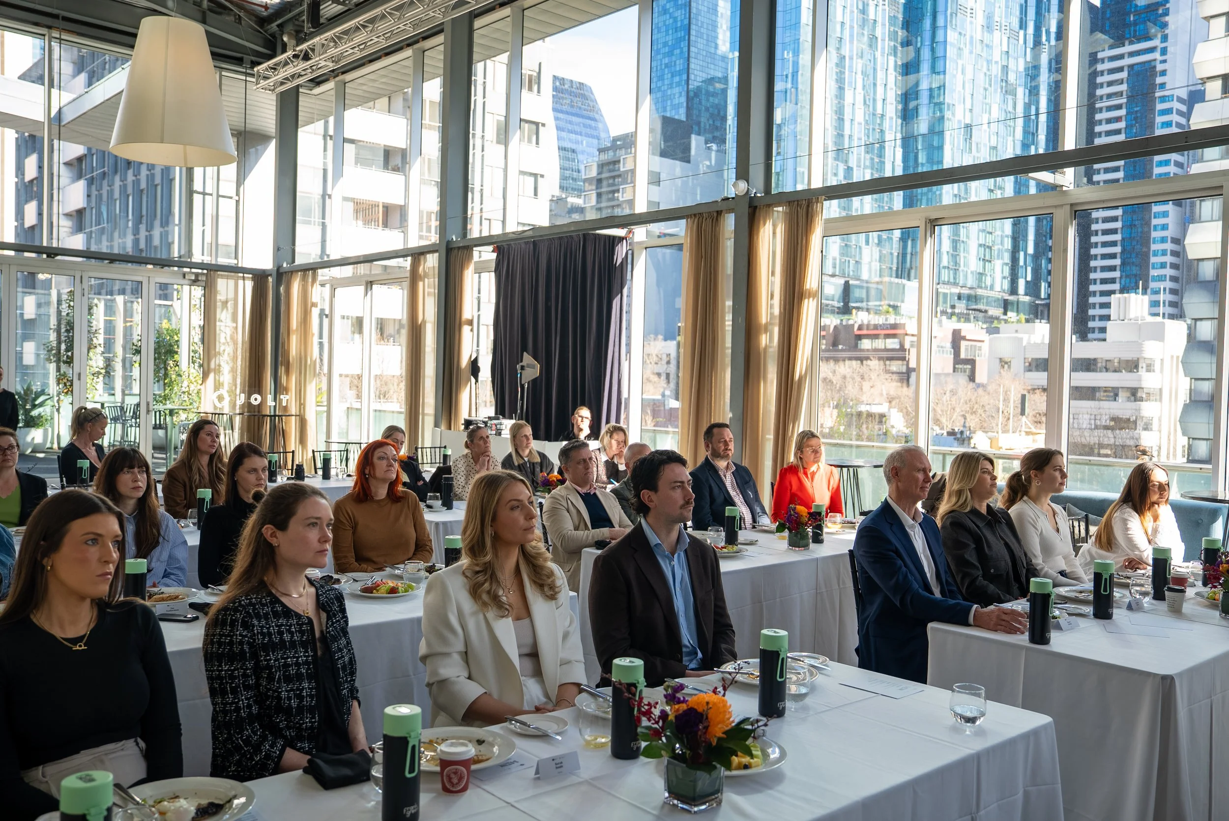 Attendees seated at round tables in a bright, modern glass-walled conference room with city skyscrapers visible outside.