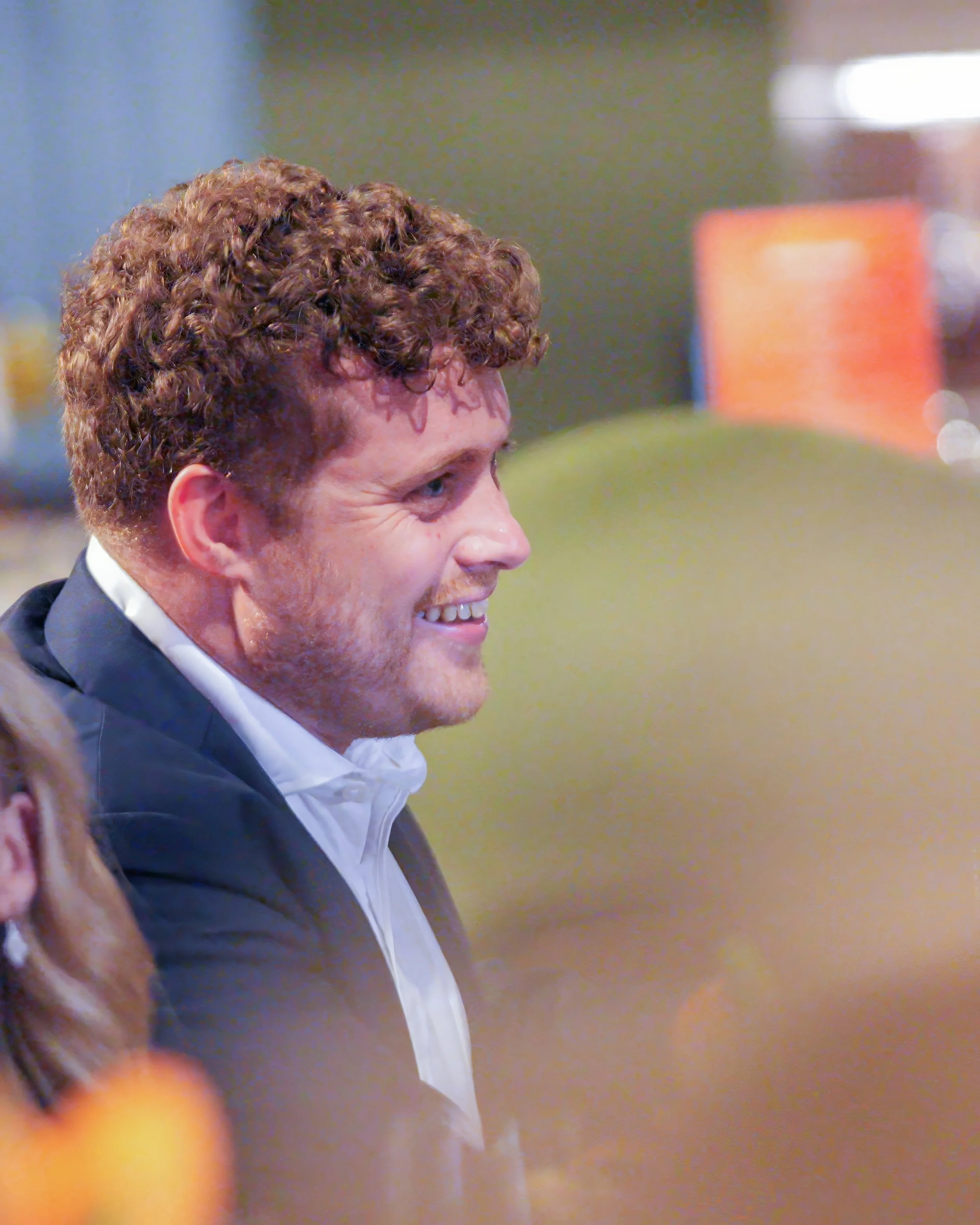 Close-up of a smiling man with curly reddish-brown hair wearing a tuxedo and white shirt at an event.