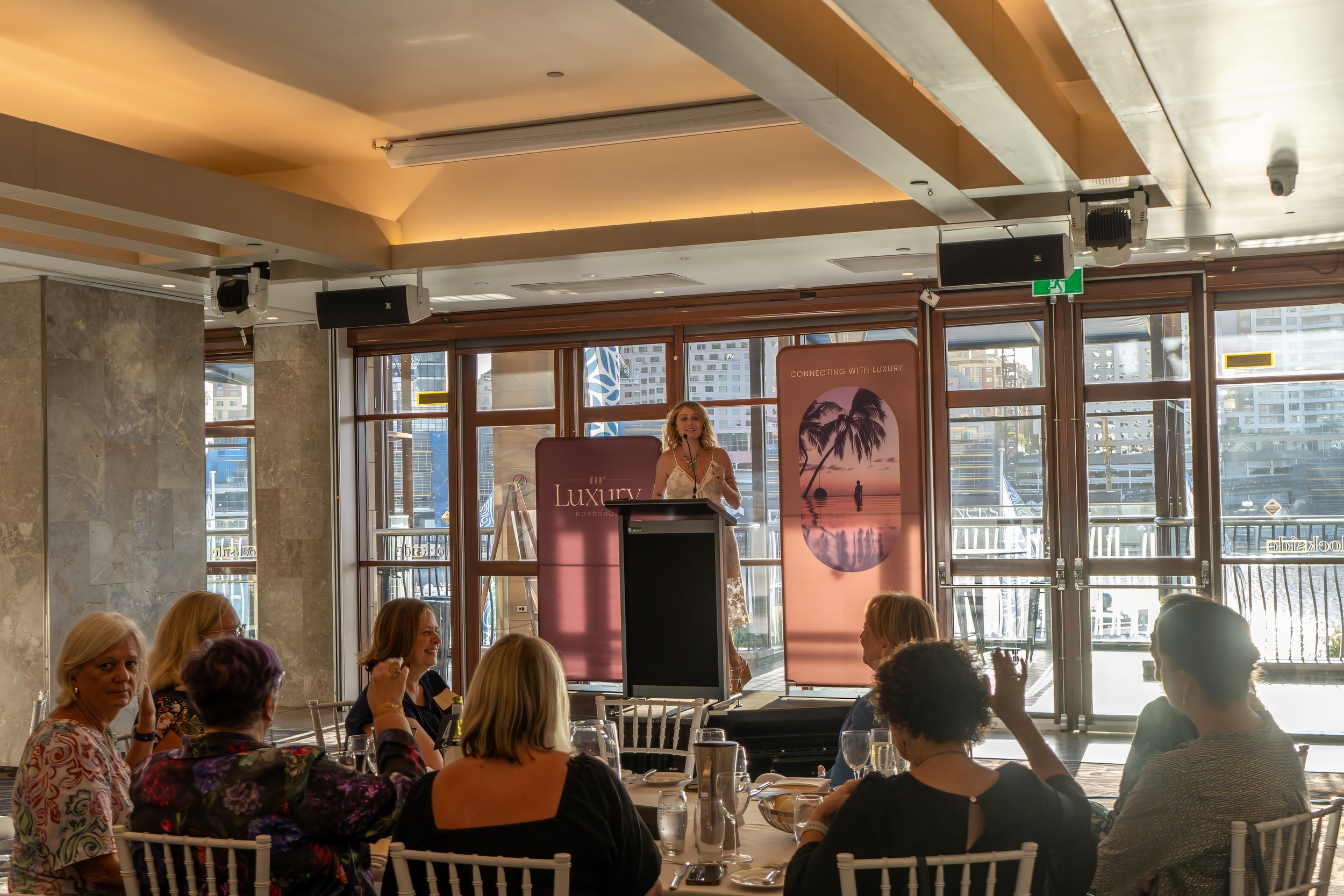 Indoor event with a woman speaking at a podium, surrounded by seated attendees, large windows with city view, promotional banners for luxury beach destination, ceiling with speakers and lighting. Brisbane. Sydney. Studios Metaversal.