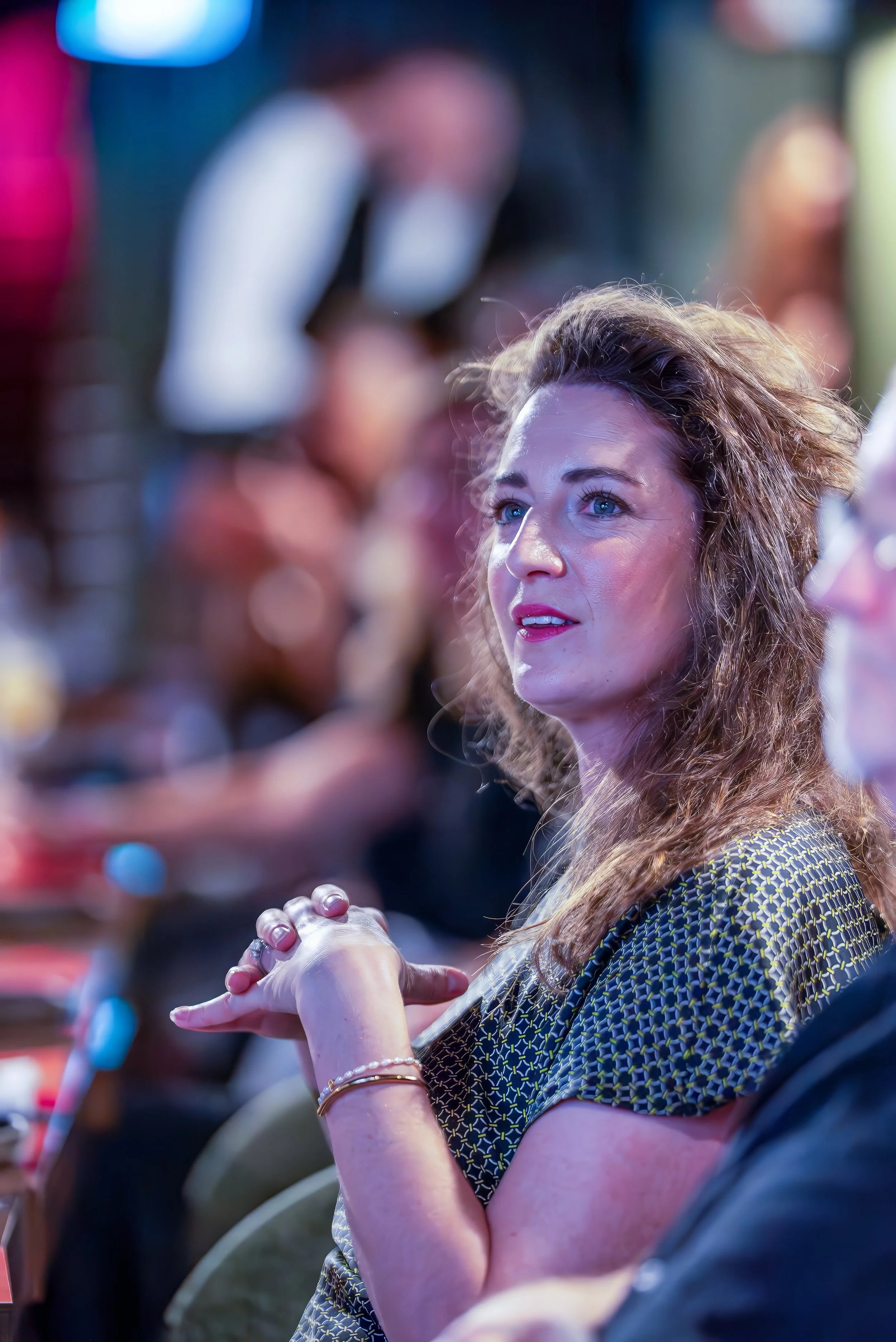 A woman with long, wavy hair and blue eyes, wearing a patterned dress and jewelry, sitting with her hands clasped together, surrounded by people at an event.