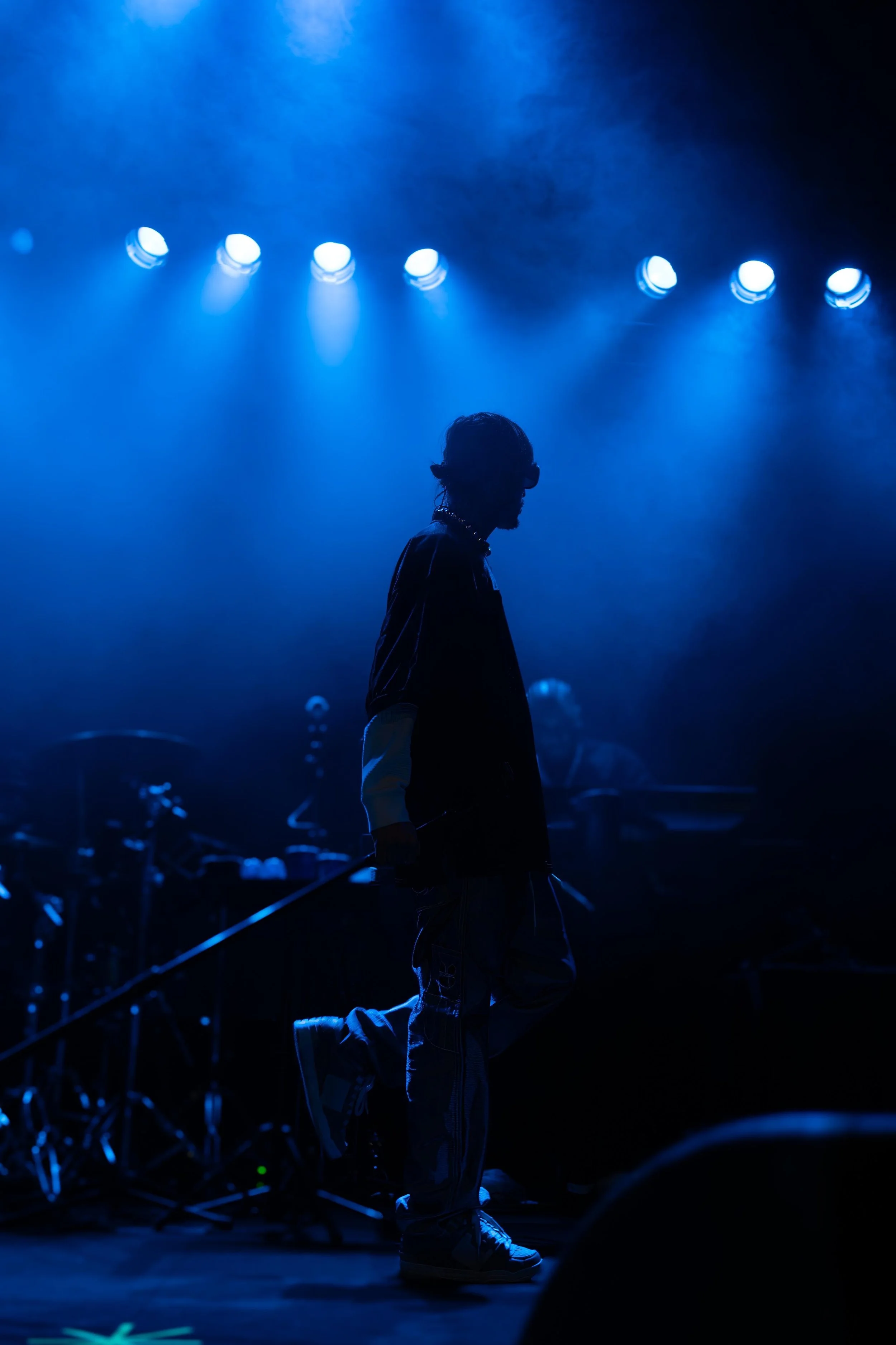 A person dressed in dark clothing with green shoes, kneeling on one knee on a stage with blue lighting, holding what appears to be a microphone, with stage lights overhead. Danny Ocean. Sydney. Studios Metaversal