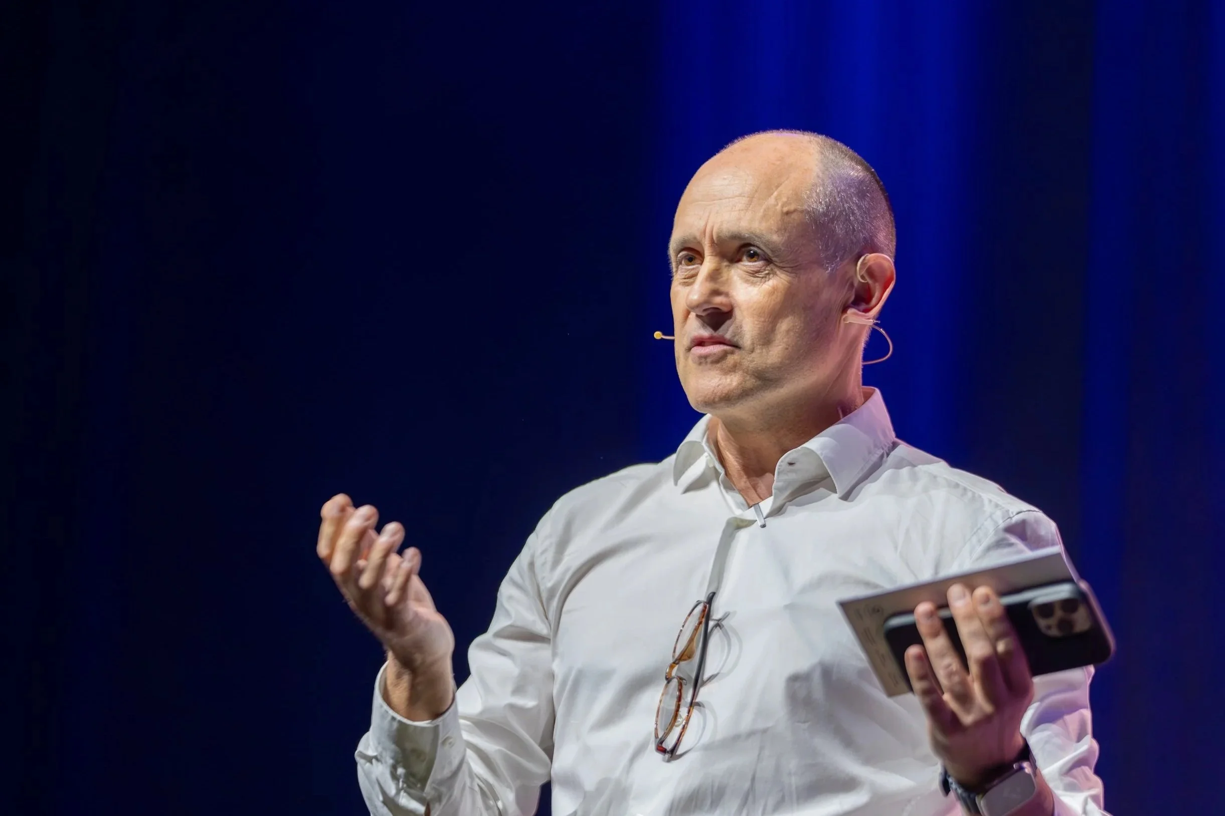 A bald man wearing a white shirt, speaking on stage with a headset microphone, holding a tablet, against a dark blue background. Sydney. Studios Metaversal