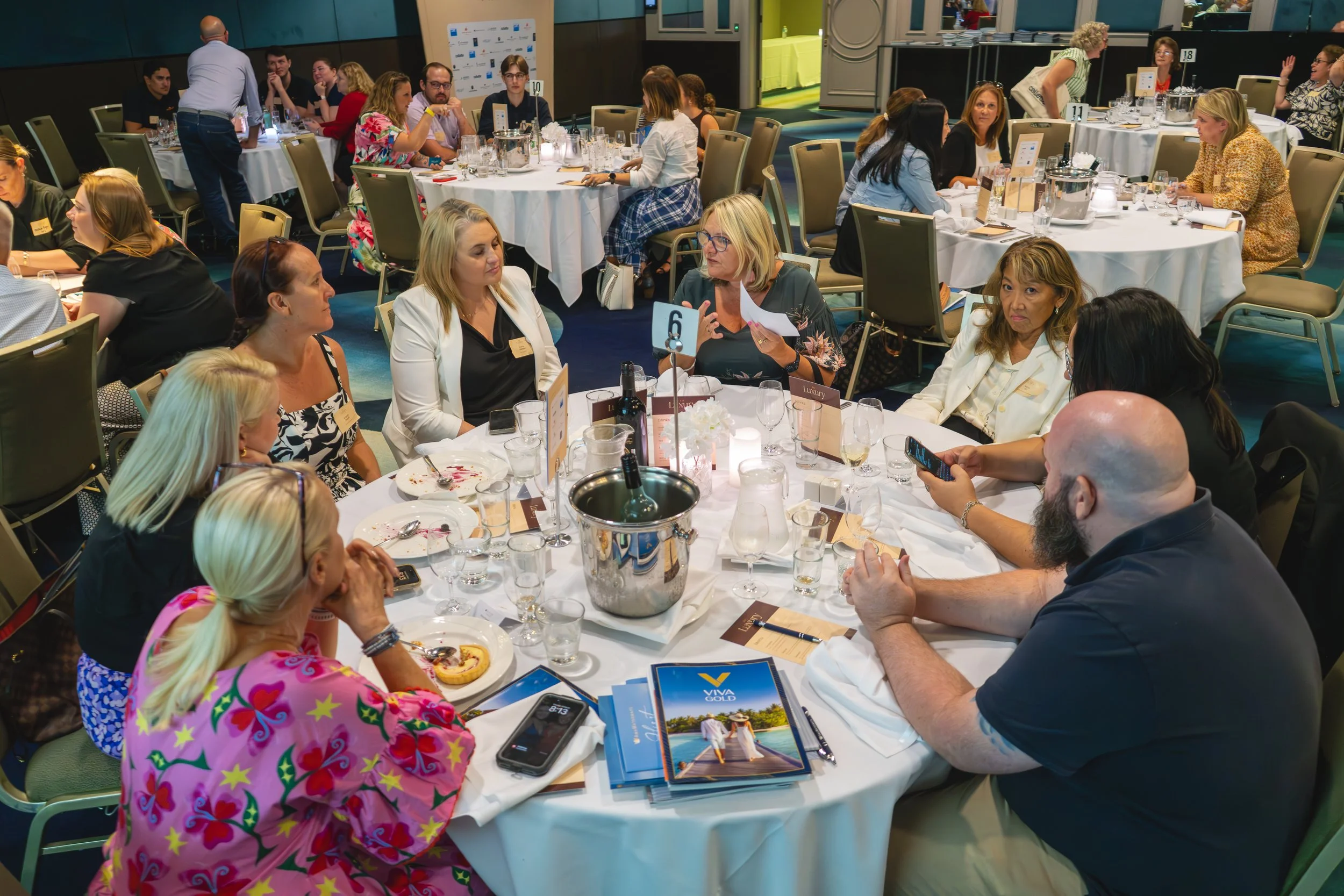 A large banquet-style room filled with round tables where people are engaged in conversations, dining, and using electronic devices. The room has a formal setting with tableware, glasses, and small table centerpieces. There are multi-generational individuals dressed in business casual and casual attire, some with name tags, indicating a professional or networking event.