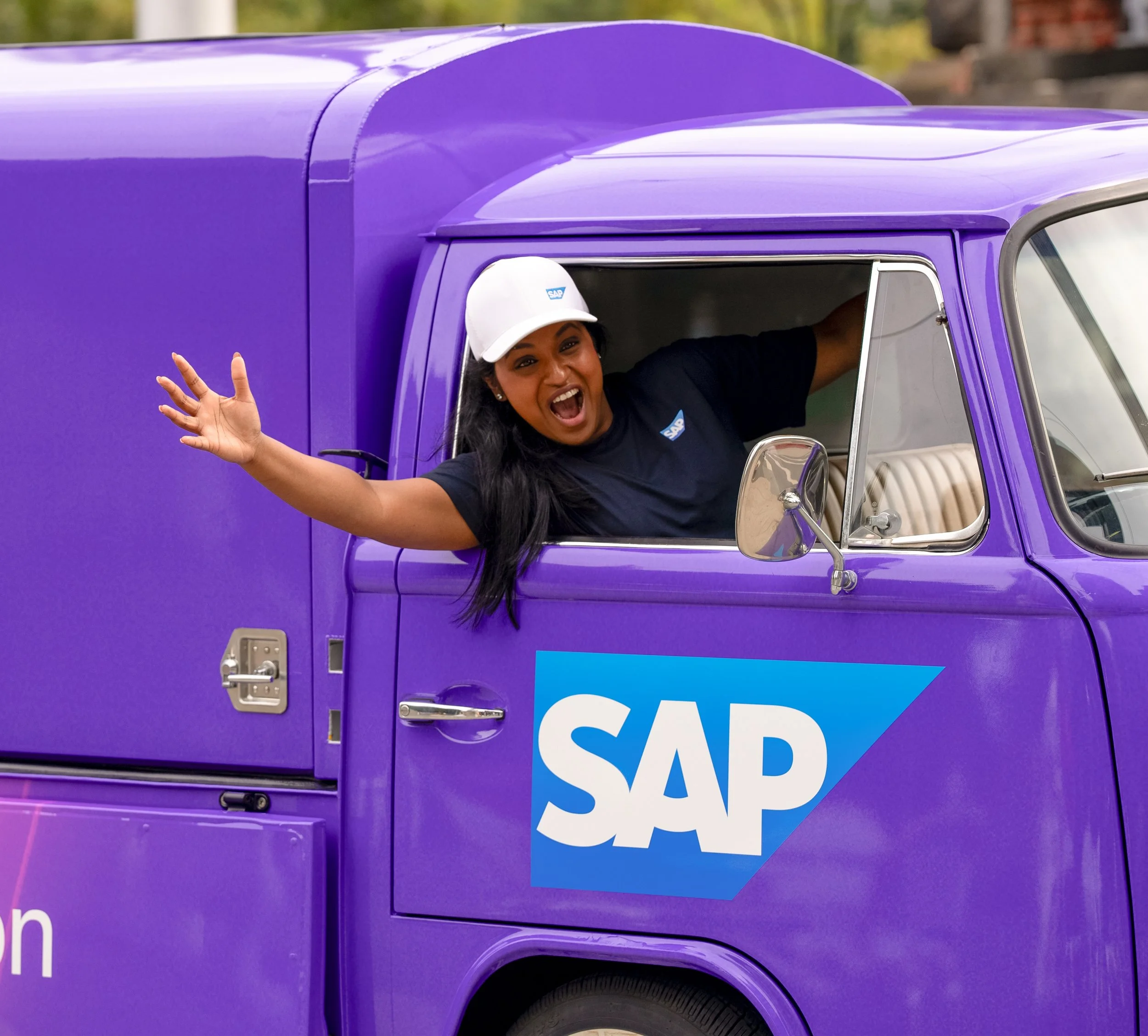 A woman smiling and waving out of the window of a purple vehicle with a partial 'SAP' logo. Melbourne. Studios Metaversal