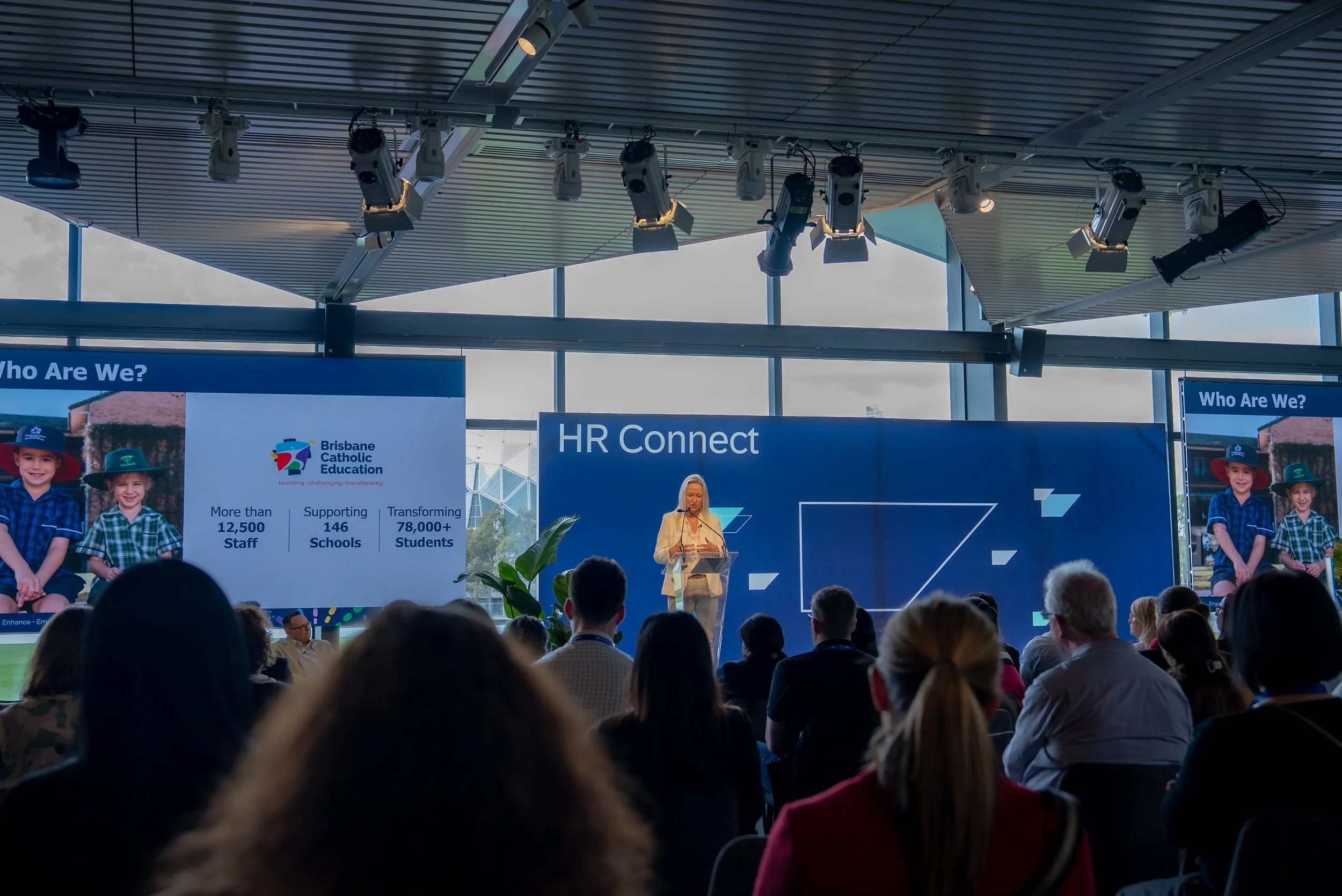 Conference or event with a woman speaking at a podium, audience seated, large screens displaying text about Brisbane Catholic Education and HR Connect, several stage lights hanging from the ceiling. SAP. Sydney. Studios Metaversal.