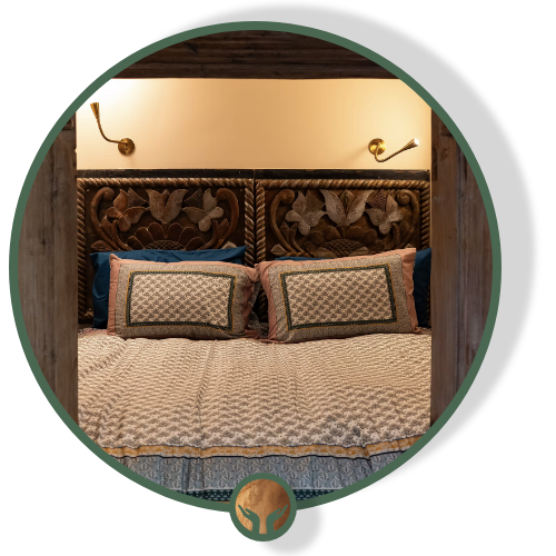 View of a bedroom bed with a carved wooden headboard, two pillows with decorative covers, and two brass wall-mounted reading lamps on either side.