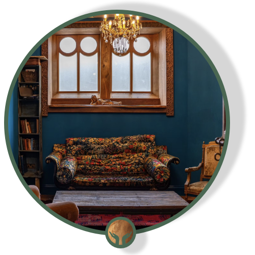 A vintage-style living room with a colorful floral-patterned sofa, wooden bookshelf, and an ornate armchair. There is a window with circular panes and a chandelier hanging from the ceiling.