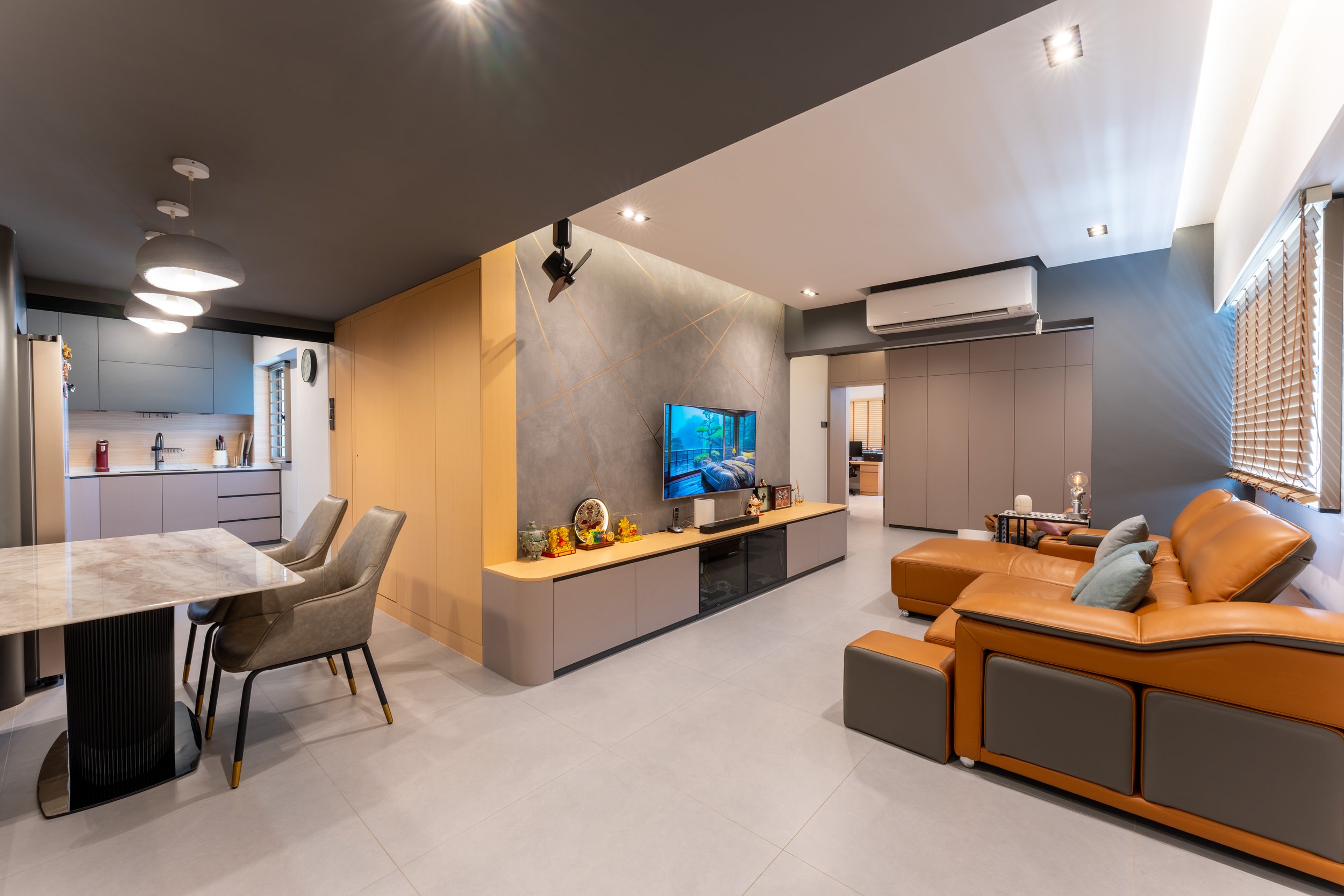 A modern living room with a beige leather sectional sofa, a wall-mounted TV, a grey wall with geometric gold lines, a wooden TV stand with traditional decorations, and a kitchen area with pendant lighting and a dining table with beige chairs.