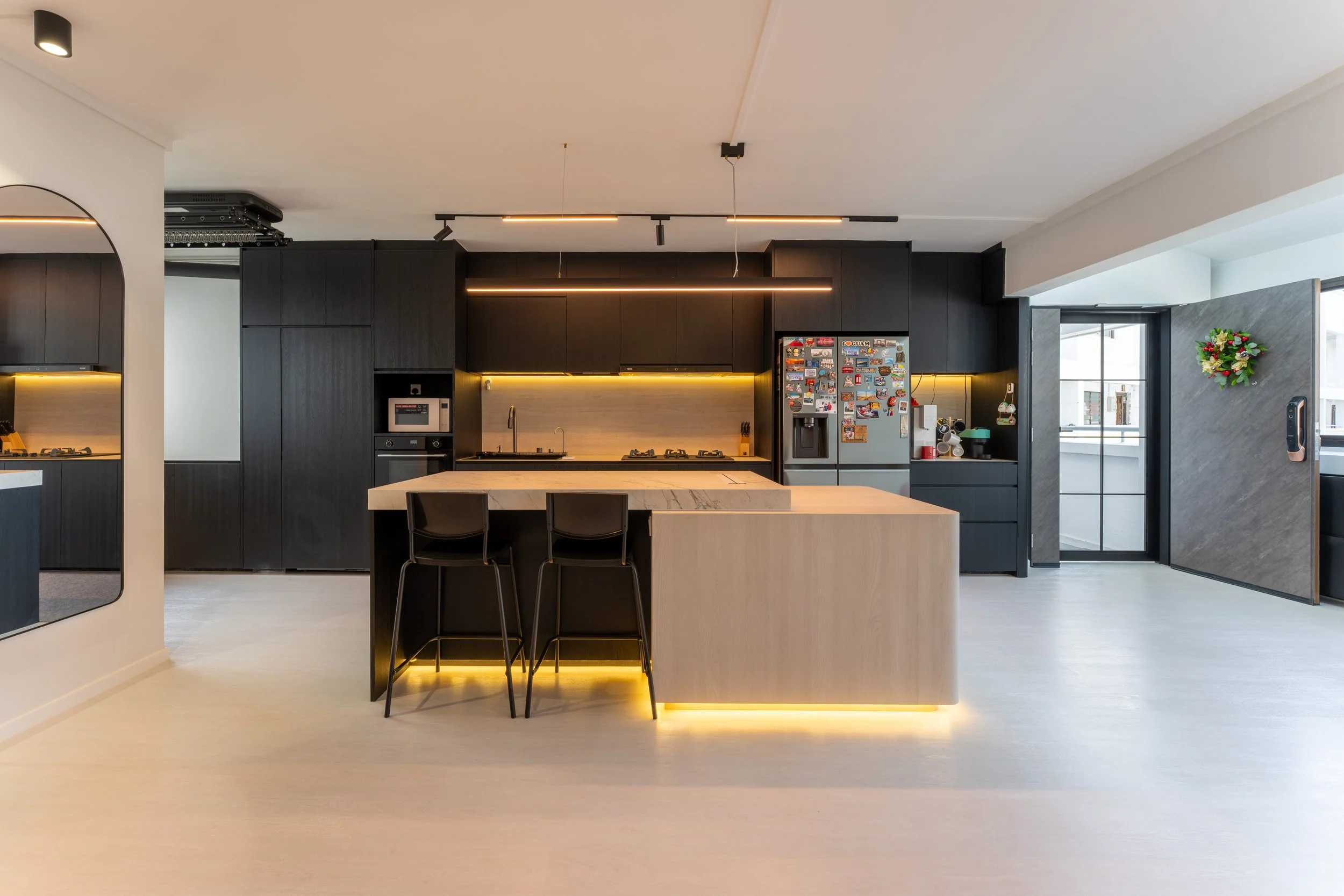 Modern kitchen with black cabinets, a white island, and a gray fridge decorated with colorful magnets. There is a small wreath on the wall near a sliding glass door, and a marble countertop with three black chairs.