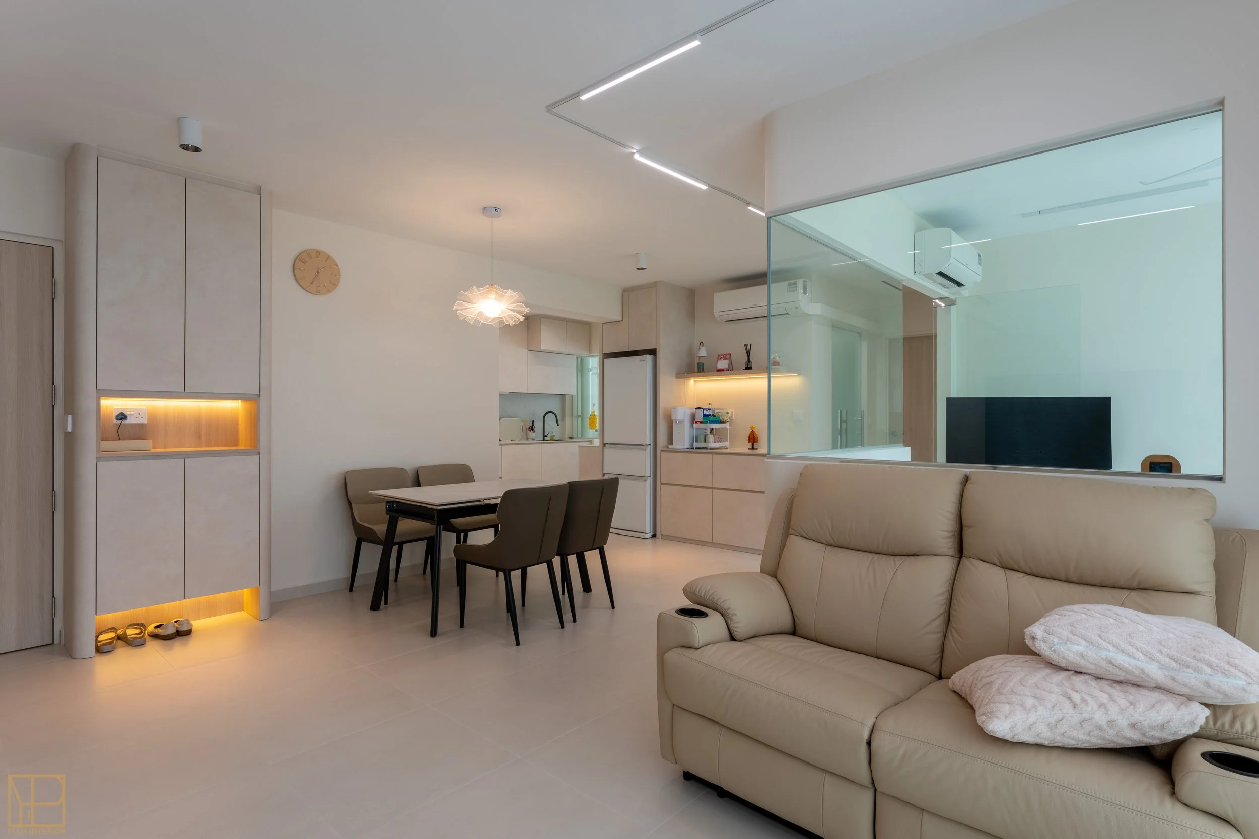 A modern, minimalistic living room and dining area with beige furniture, a round wall clock, a kitchen with white cabinetry and a fridge, a glass partition, and contemporary lighting fixtures.