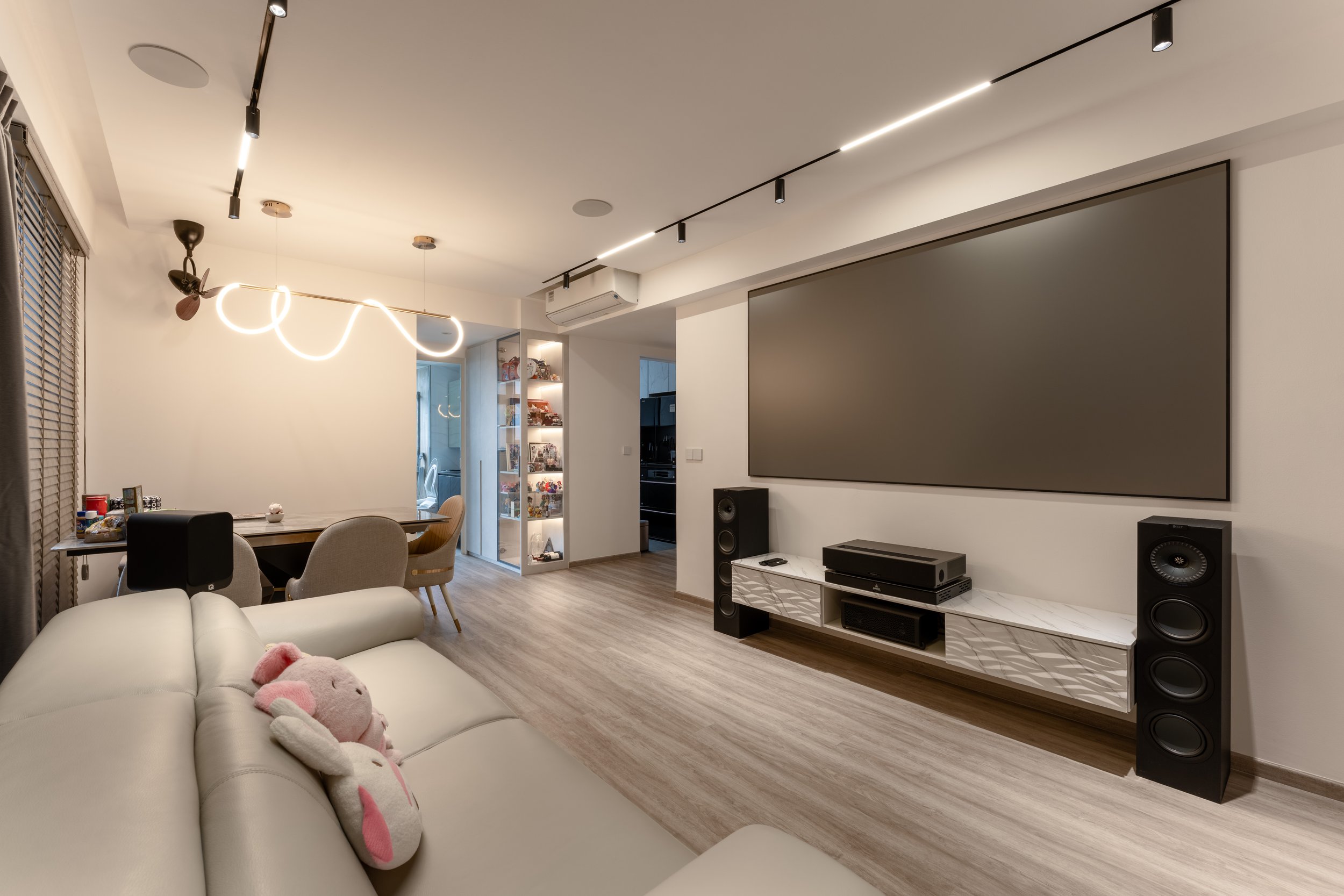 Modern living room with a white sofa, pink plush rabbit toy, large flat screen TV, wood flooring, ceiling lights, and a decorative display cabinet.
