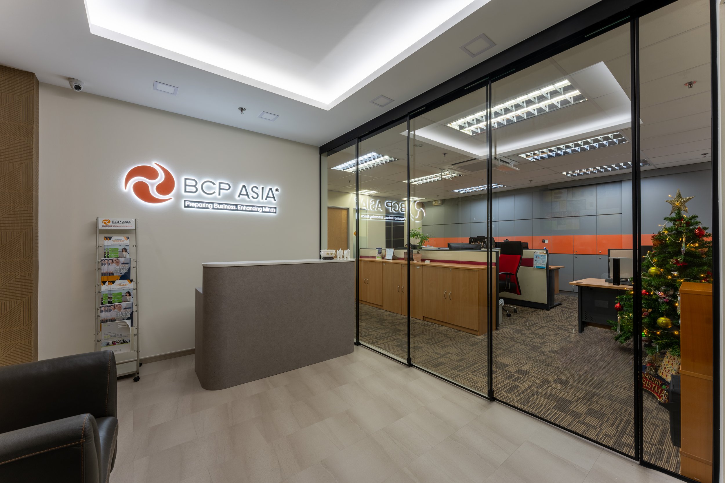 Reception area of BCP Asia office with a Christmas tree, a curved gray reception desk, and a glass wall behind the desk. The office has wood and gray decor, and the company's logo is illuminated on the wall.
