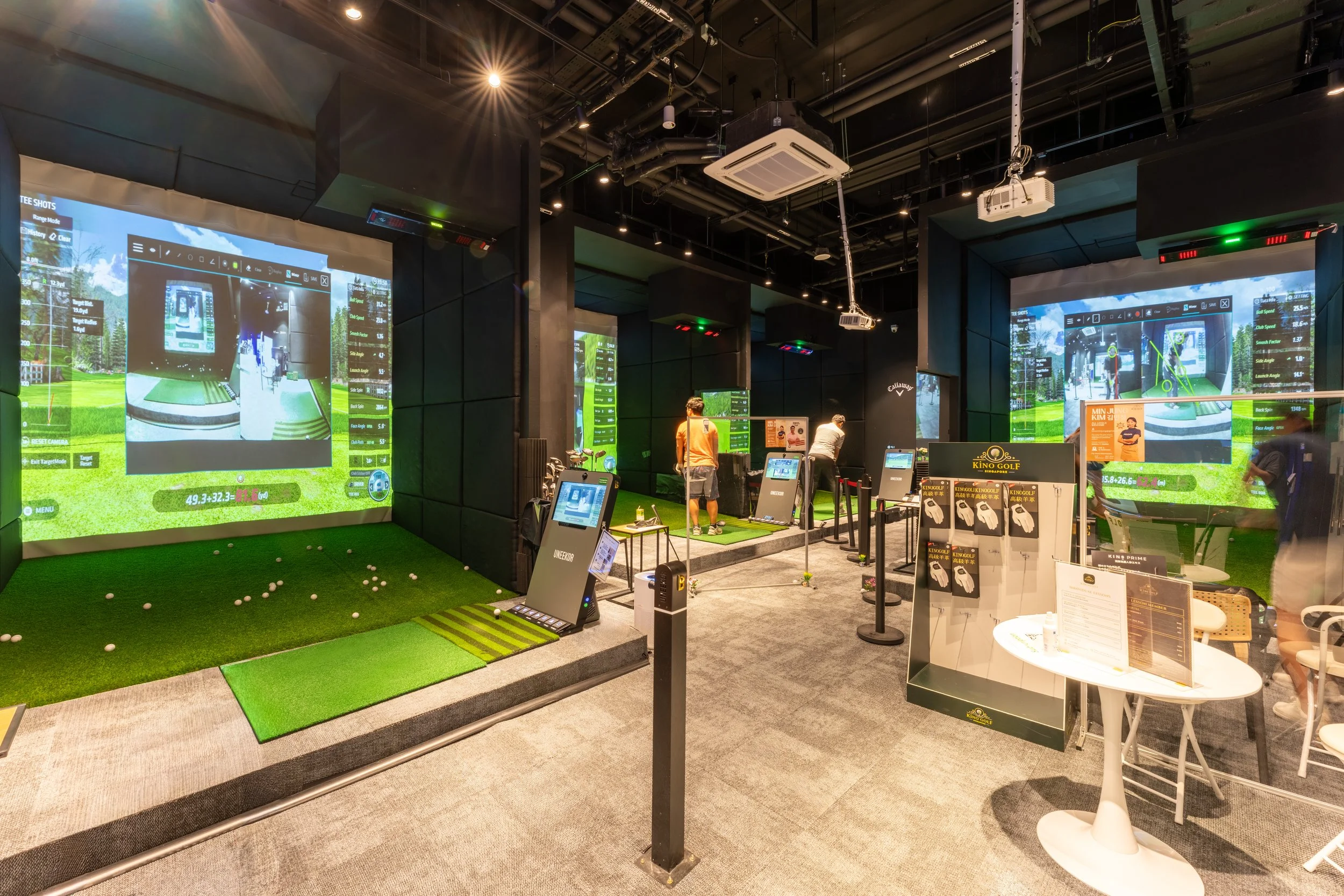 Indoor golf simulation room with multiple screens showing golf course images, golf equipment, and two people practicing their swings. There are golf nets, balls, and simulation technology visible.